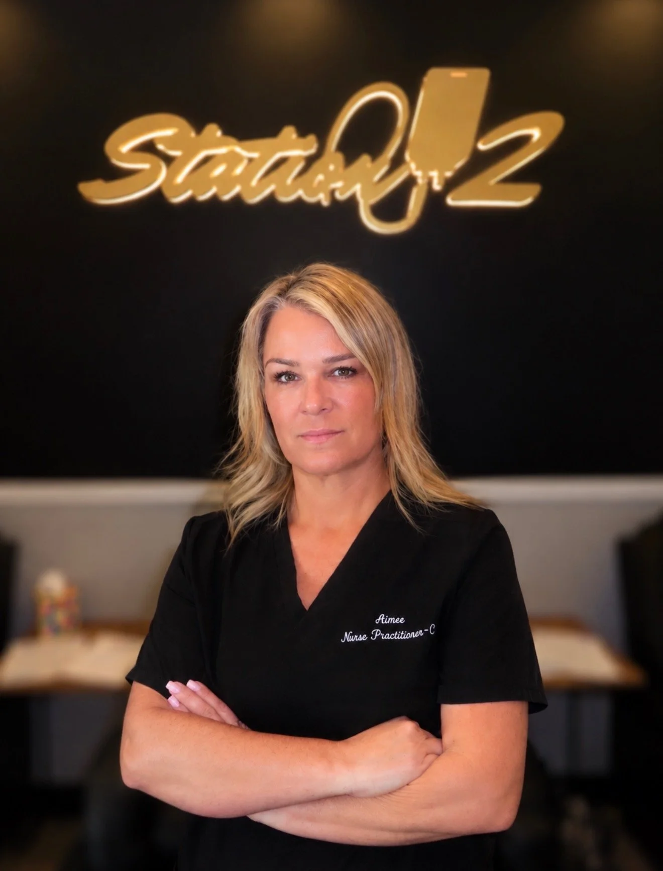 A woman with blonde hair, wearing a black nurse uniform with her arms crossed, standing in front of a black wall with illuminated gold sign that reads 'Startup' and '2'.