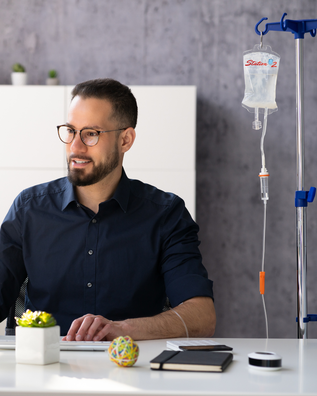 A man with glasses and a beard sitting at a desk with a laptop, a notebook, a plant, and a colorful rubber band ball, with an IV drip bag hanging nearby.