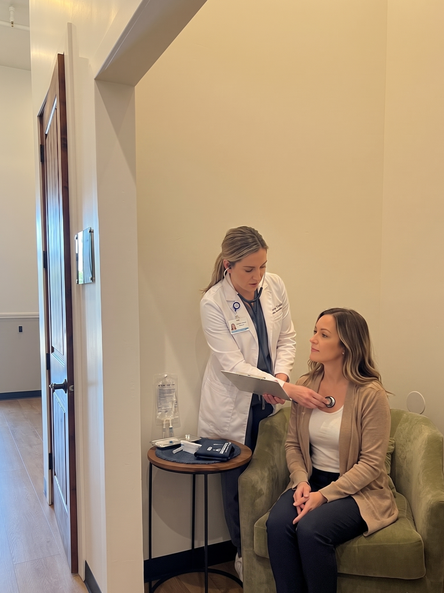A healthcare professional, possibly a nurse or doctor, examining a woman seated on a green couch in a medical office. The professional is holding a clipboard and using a stethoscope on the patient's chest.