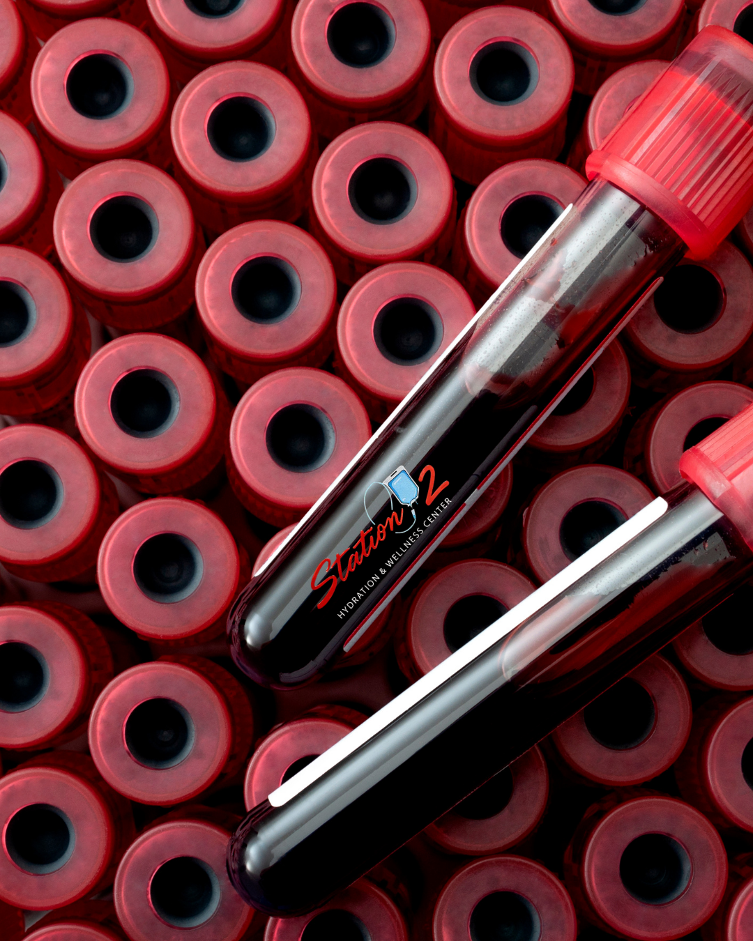 Close-up of multiple blood sample test tubes with red caps, arranged in a grid. Two test tubes in the foreground contain blood samples; one has a label reading 'Station 2 Hydration & Wellness Center'.