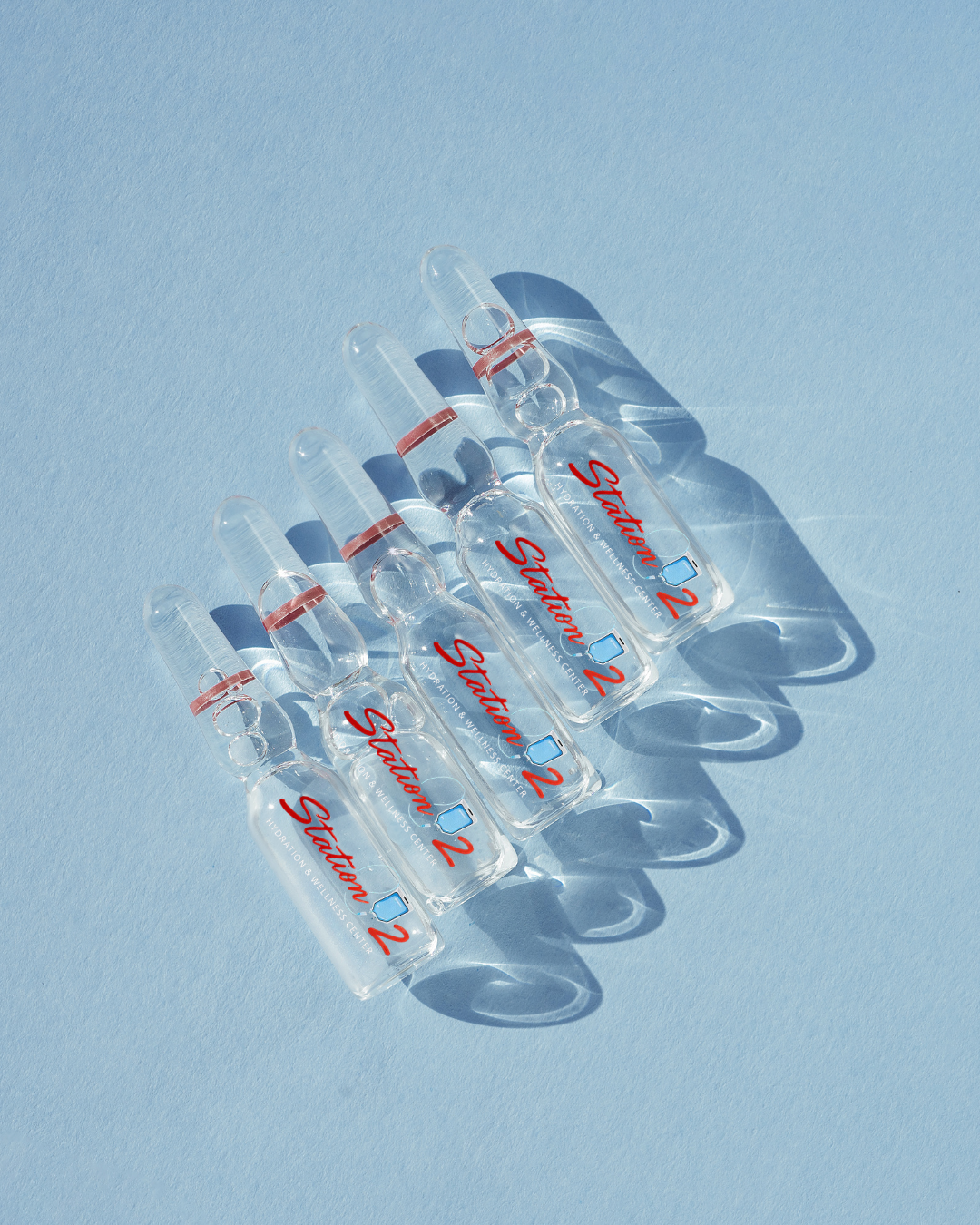 Five small glass bottles labeled in red with blue accents, arranged on a light blue surface, casting shadows.