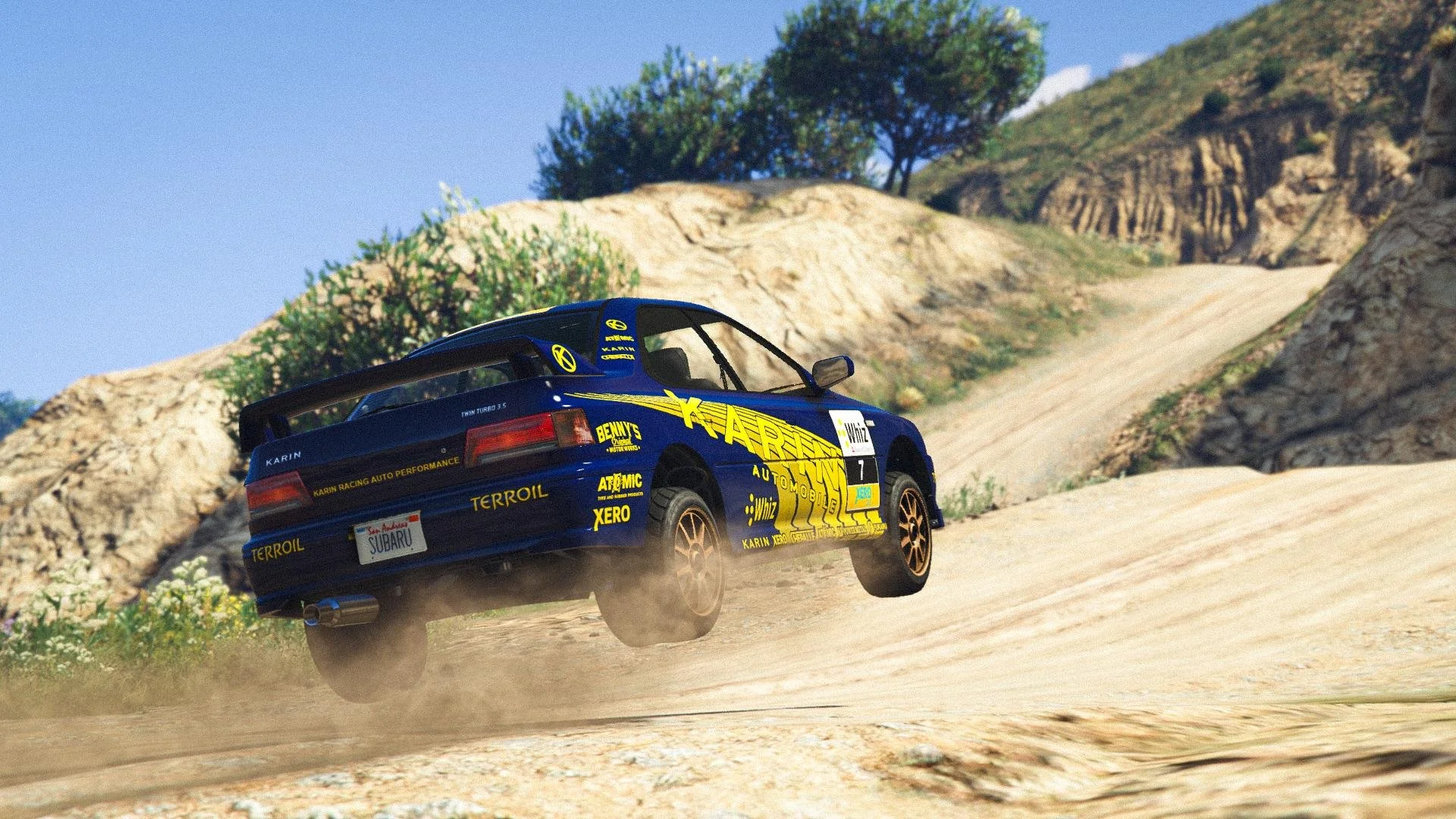 A blue rally car with yellow and black sponsor decals speeds on a dusty dirt road in a mountainous area with green bushes and rocky terrain.