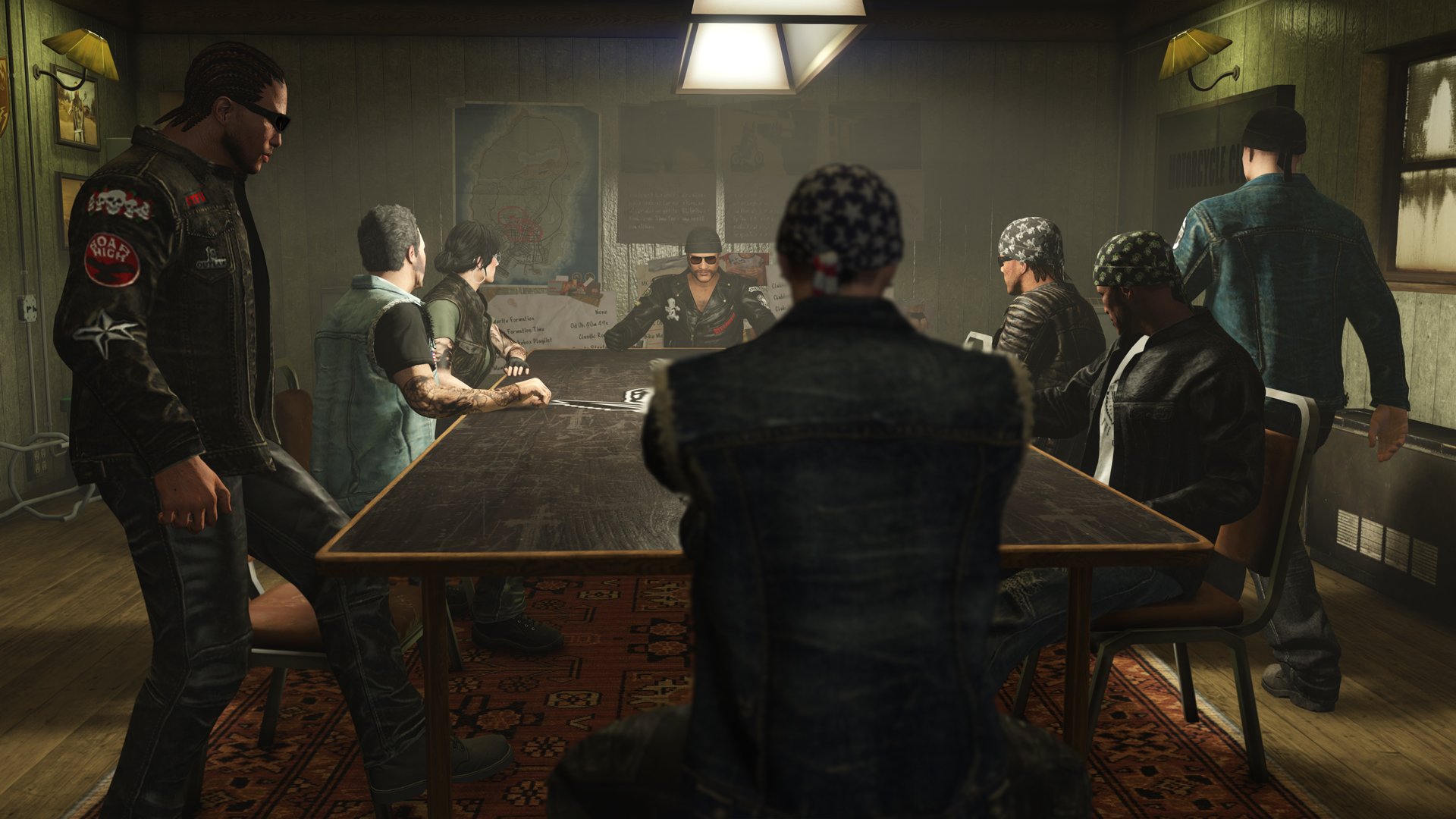 A group of people dressed in biker and gang-style clothing are sitting and standing around a large conference table in a dimly lit room, with one person at the head of the table appearing to lead the discussion.