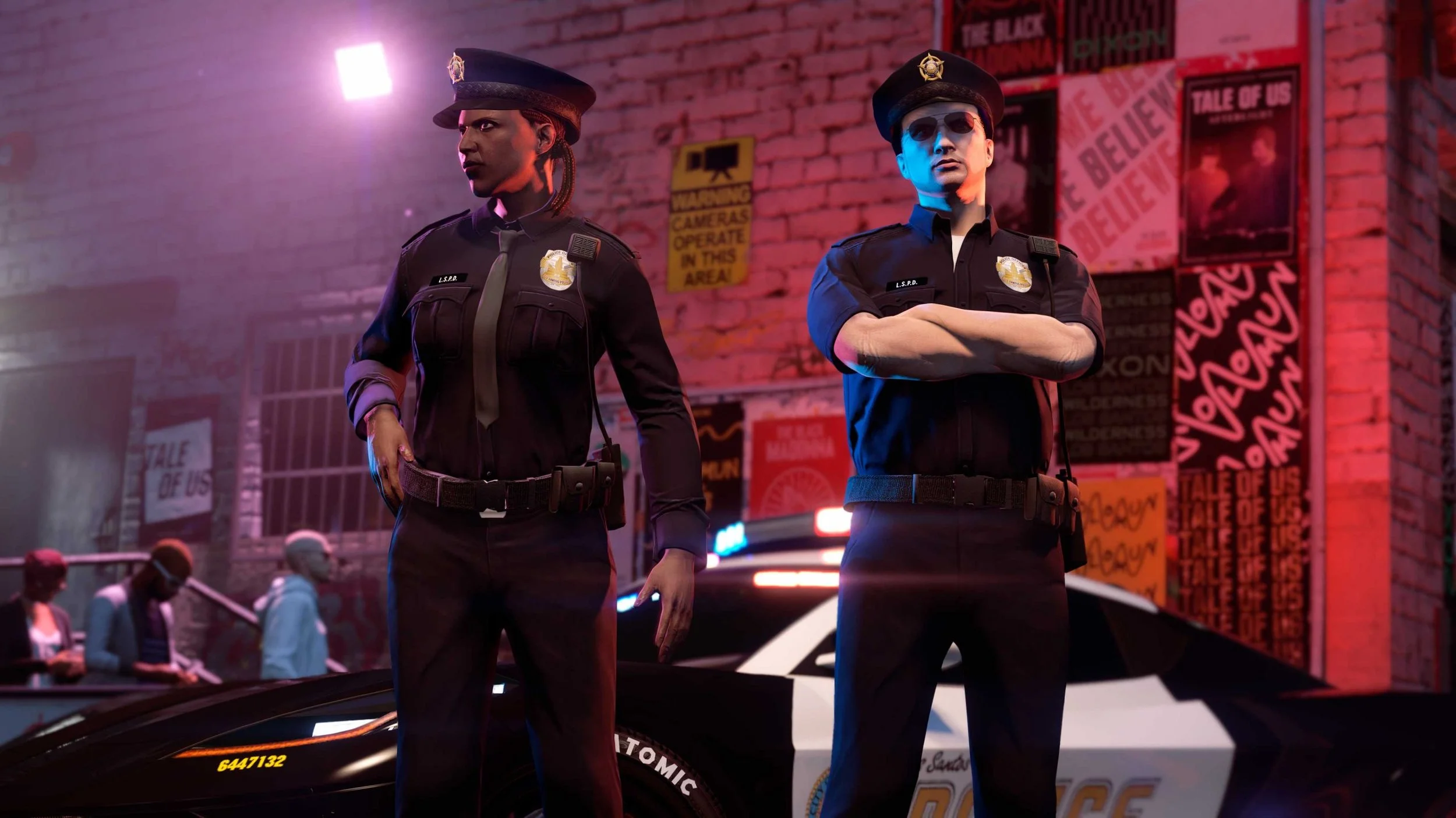 Two police officers in uniform standing with arms crossed in front of a brick wall with various posters and signs, including one that reads 'TALE OF US' and another that says 'Caution Cameras Operate in This Area,' in a nighttime urban setting.