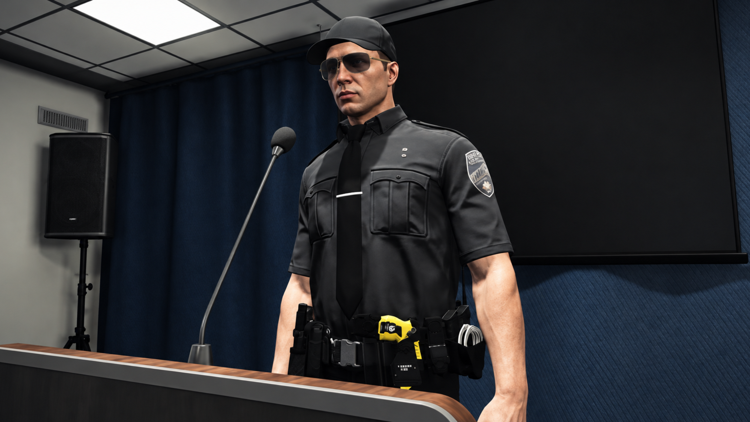 A man dressed as a police officer standing behind a table, wearing sunglasses, a police uniform, and a black cap, with a microphone on a stand, a speaker, and a large monitor in the background in a conference room.