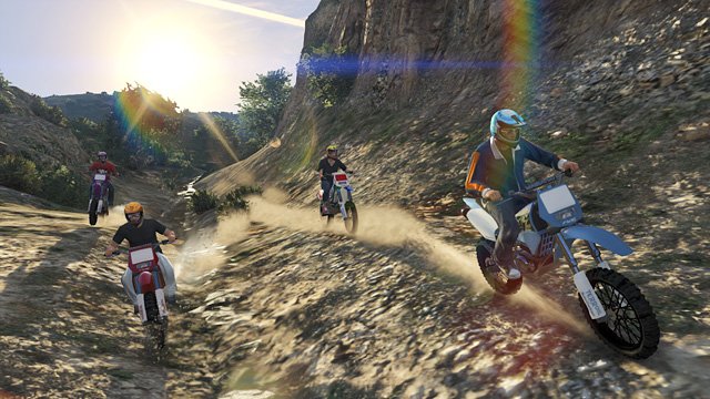 Four dirt bike riders, wearing helmets, riding through a rocky off-road trail with dust and a dramatic landscape, under a sunny sky with rainbow flares.