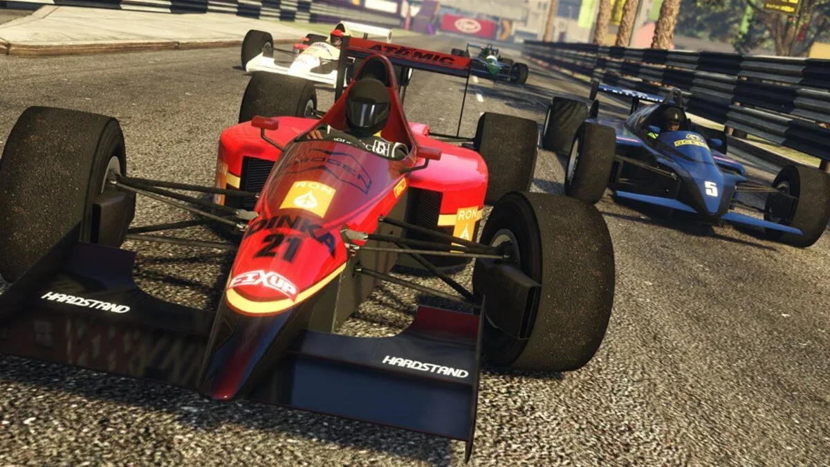 Several open-wheel racing cars on a street circuit track, with the car in the foreground being red numbered 21, and others behind in various colors, racing closely together.
