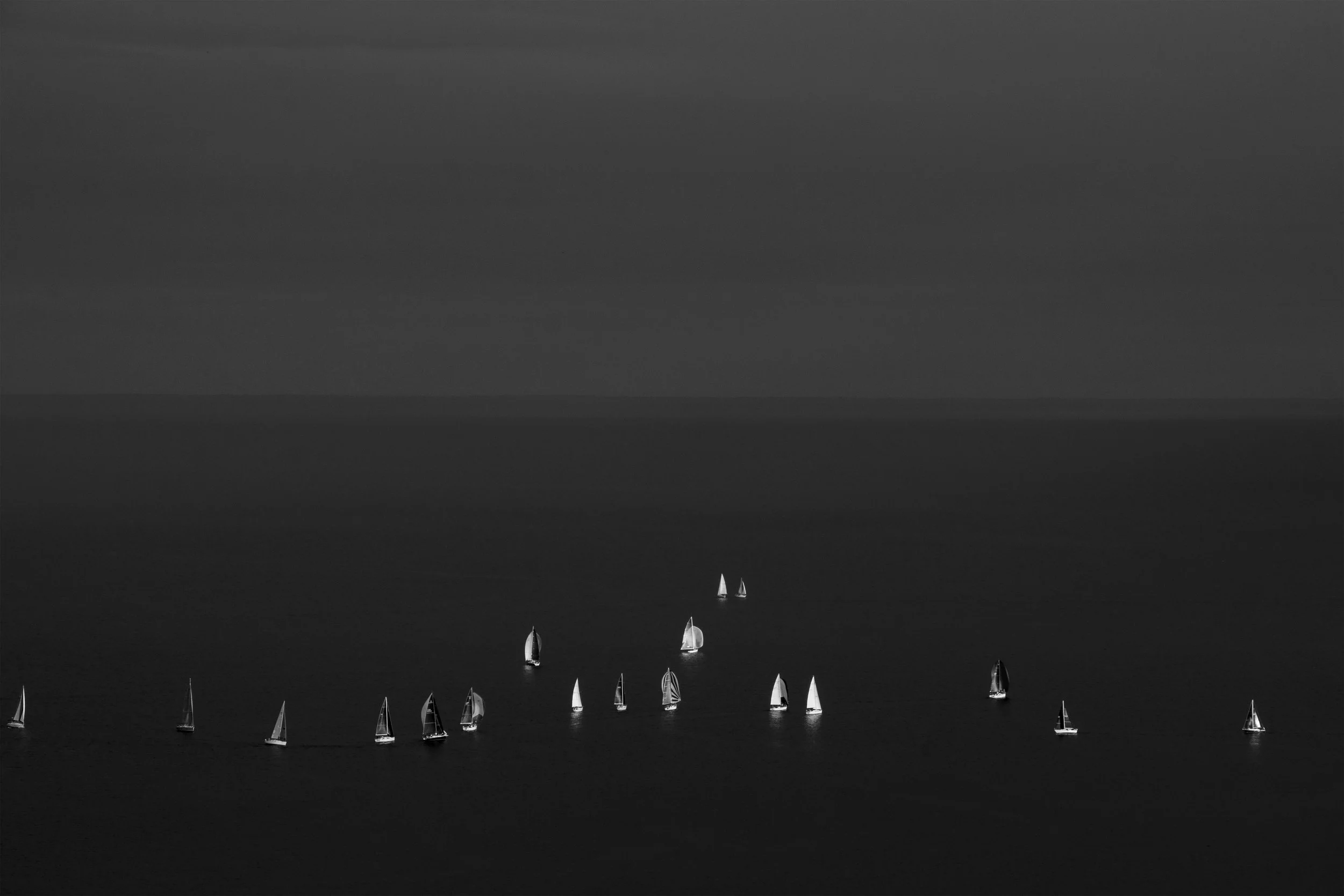 Black and white photograph of multiple sailboats on the water, with a calm horizon and cloudy sky in the background.