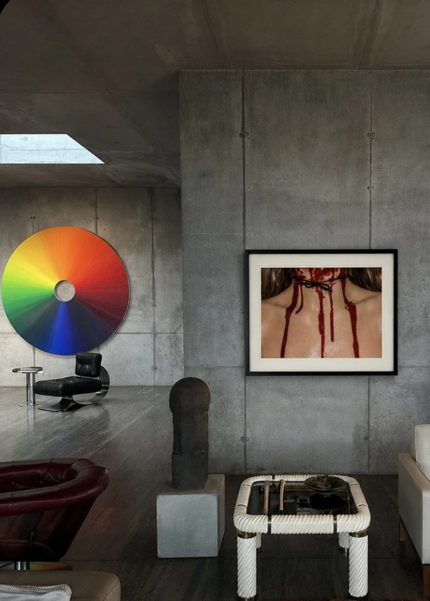 Interior of a modern art gallery with a concrete wall. On the wall, there is a framed photo of a woman's neck with blood dripping from a wound. To the left, there is a large color wheel and a black modern chair.