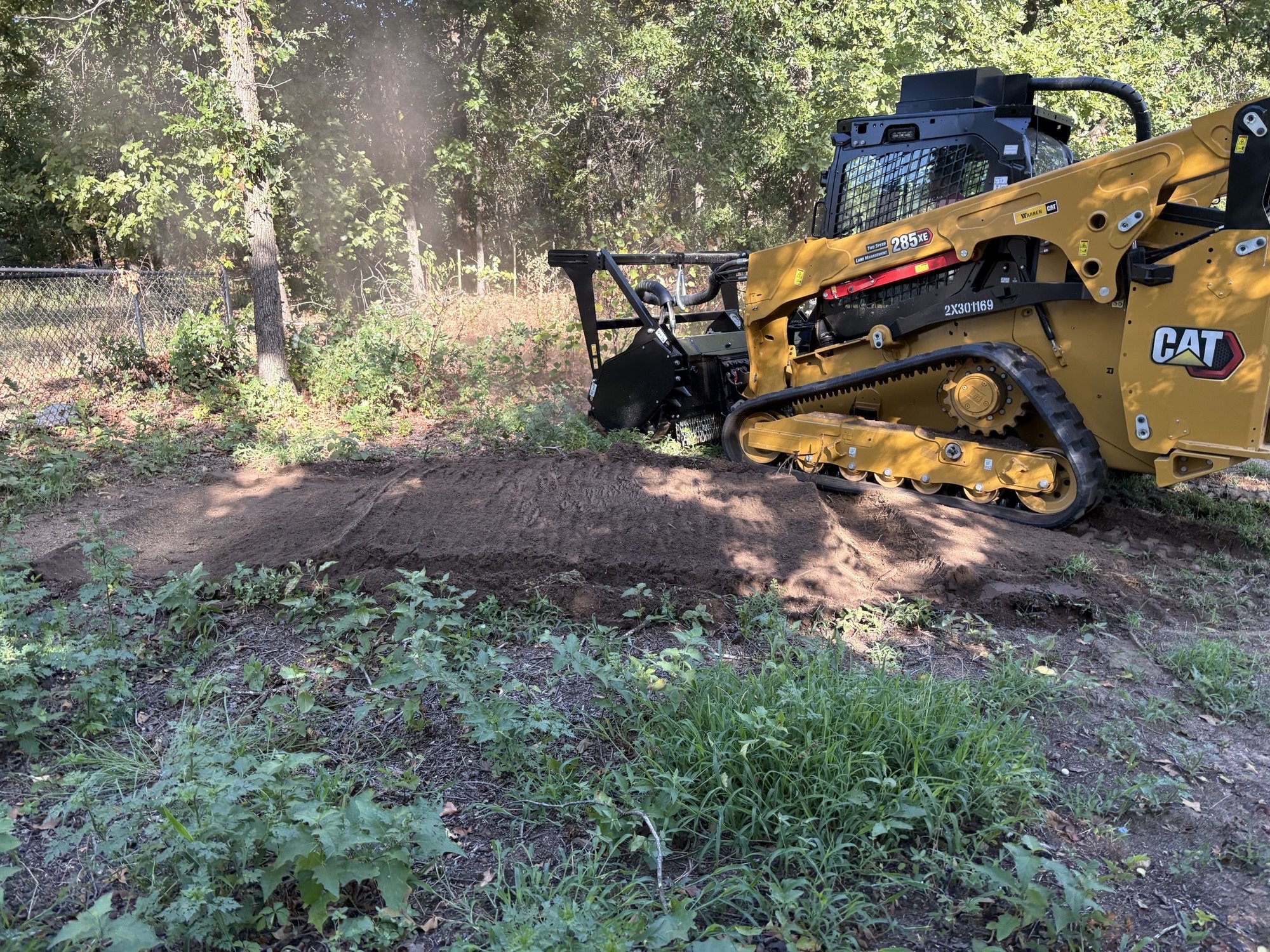 A yellow compact excavator operating on a dirt and grassy area near a forested area with a chain-link fence in the background.