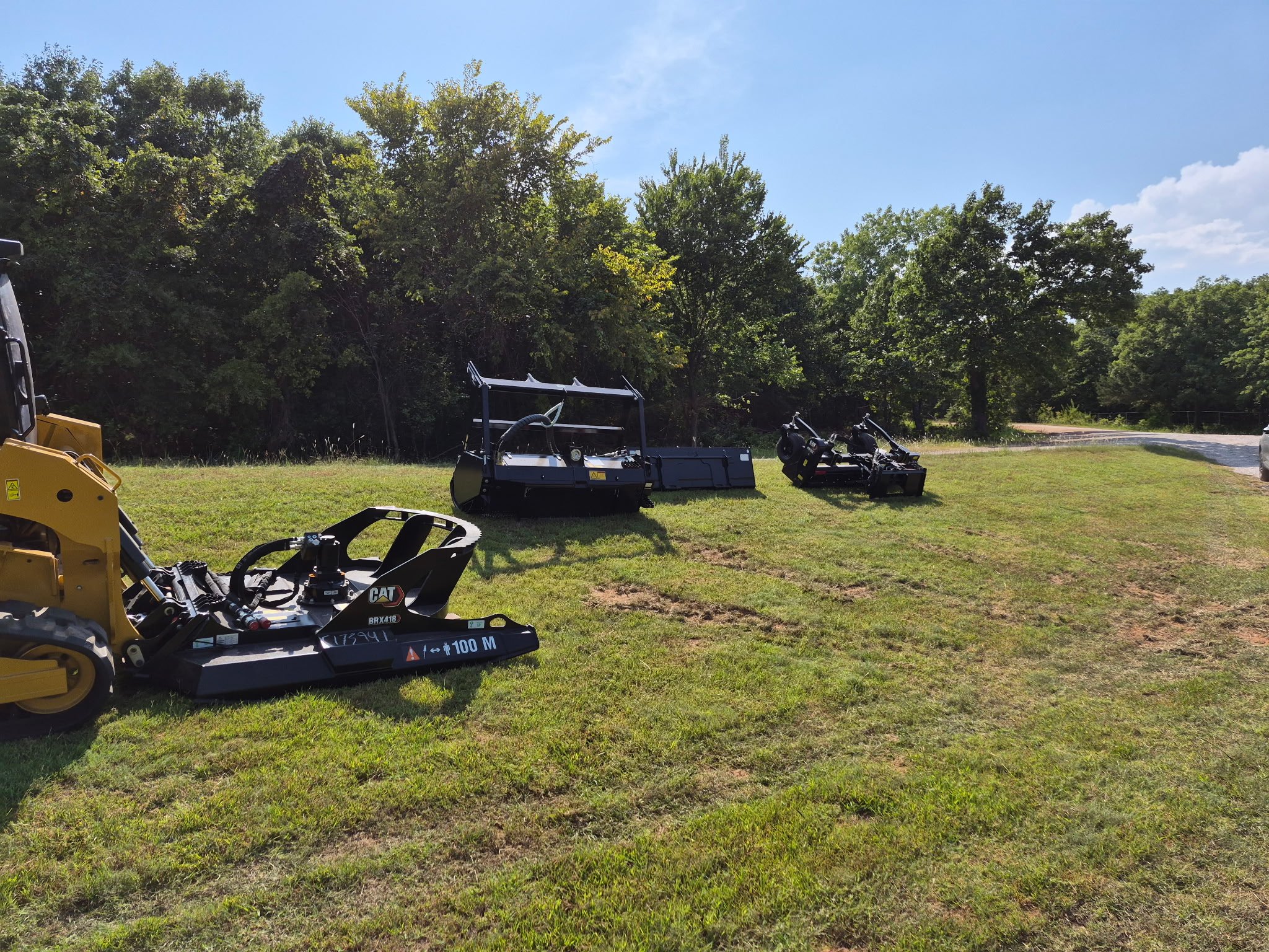 Various pieces of landscaping or construction equipment and machinery on a grassy area with trees and blue sky in the background.