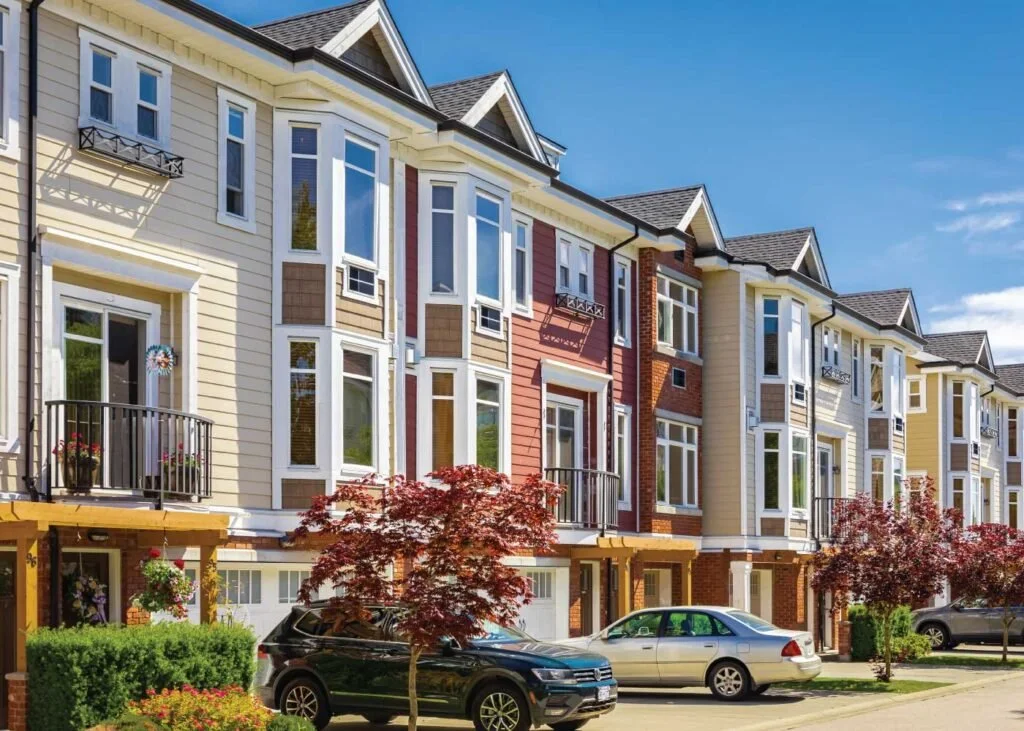 Row of modern townhouses with bay windows and small balconies, parked cars, trees, and a bright blue sky.