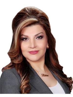 A woman with long brown hair styled in loose curls, wearing a gray blazer and white top, with the name 'Binna' on a necklace, posing for a portrait against a plain white background.