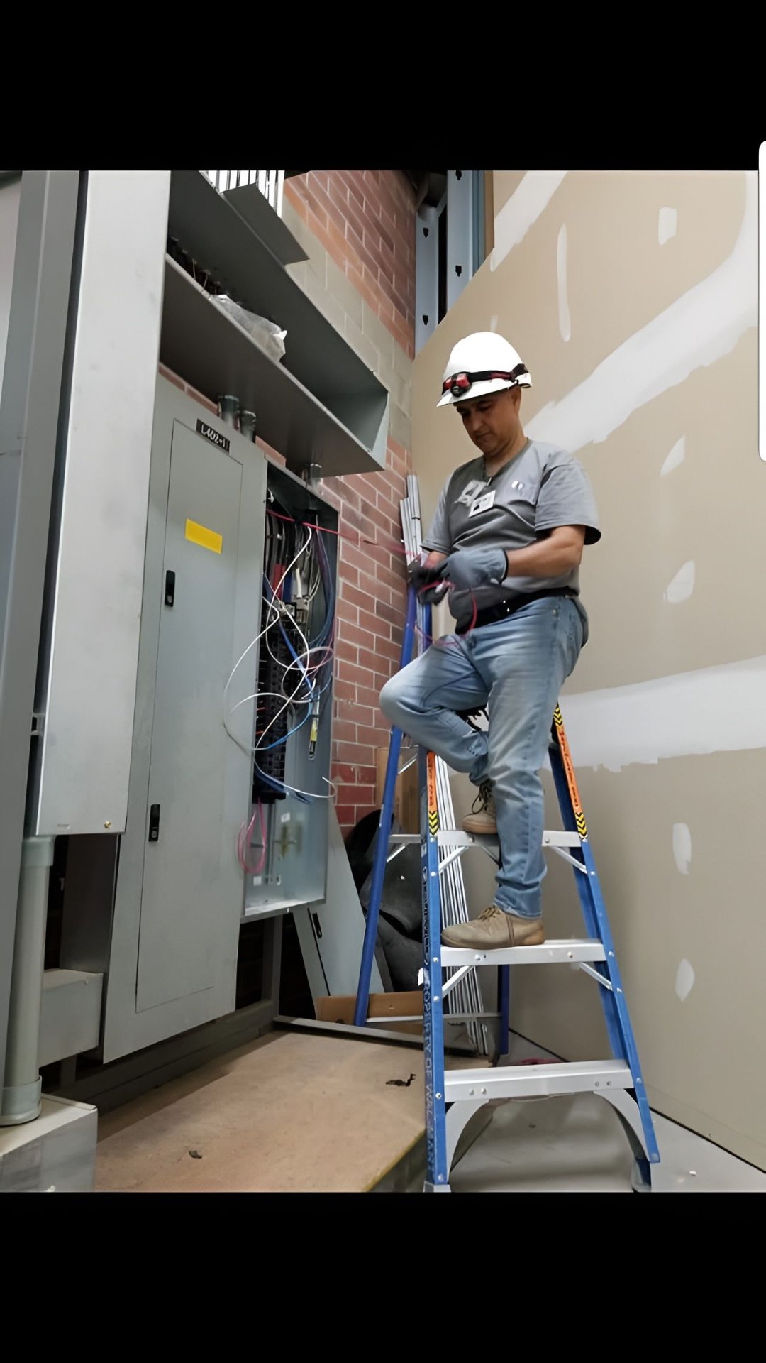 A worker wearing a white safety helmet, gray shirt, and jeans is standing on a blue step ladder, working on an electrical panel. The background features a brick wall and unfinished drywall.