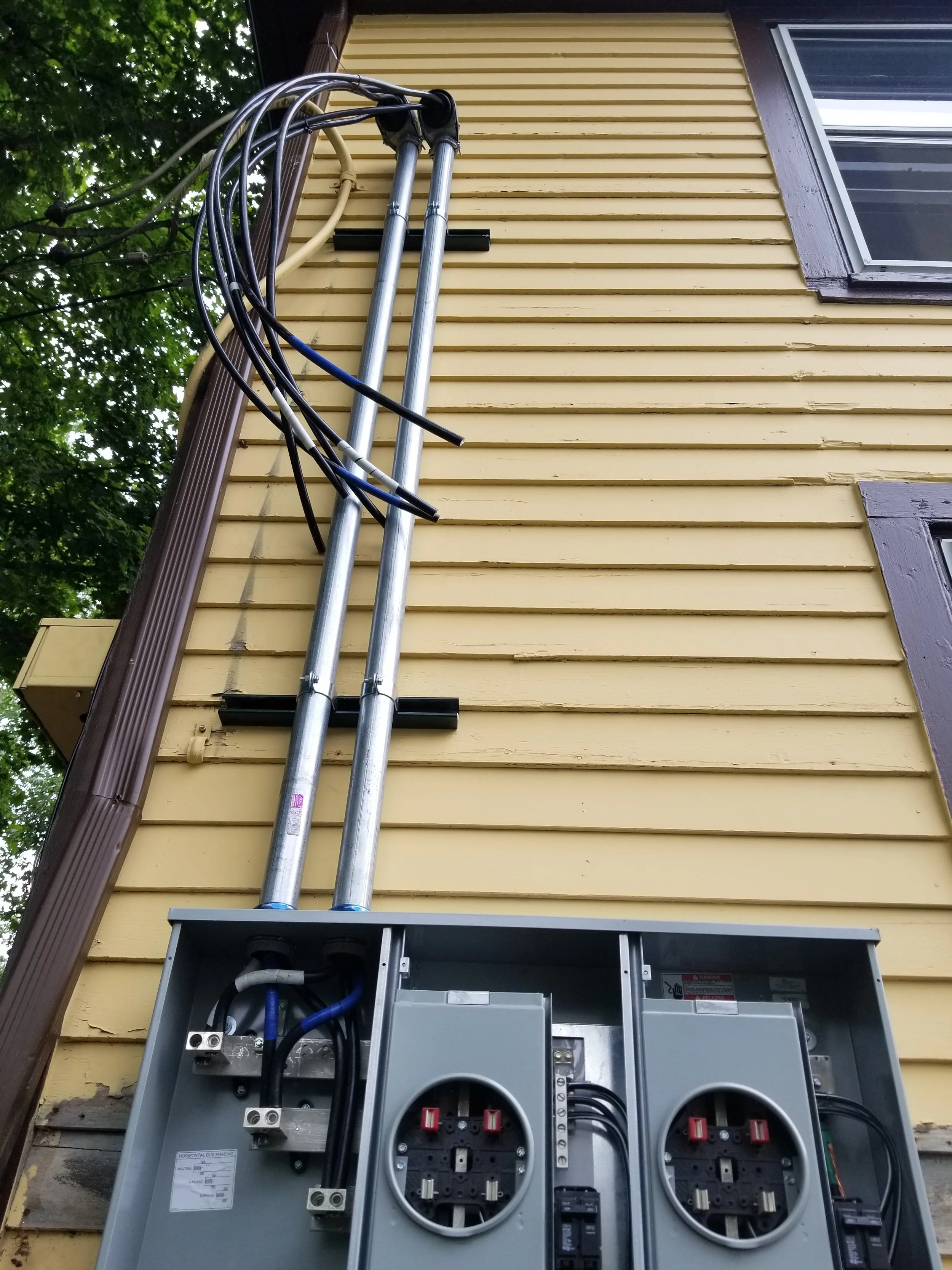 Electrical breaker panel with wires and conduits mounted on the exterior of a yellow house with dark brown trimming.