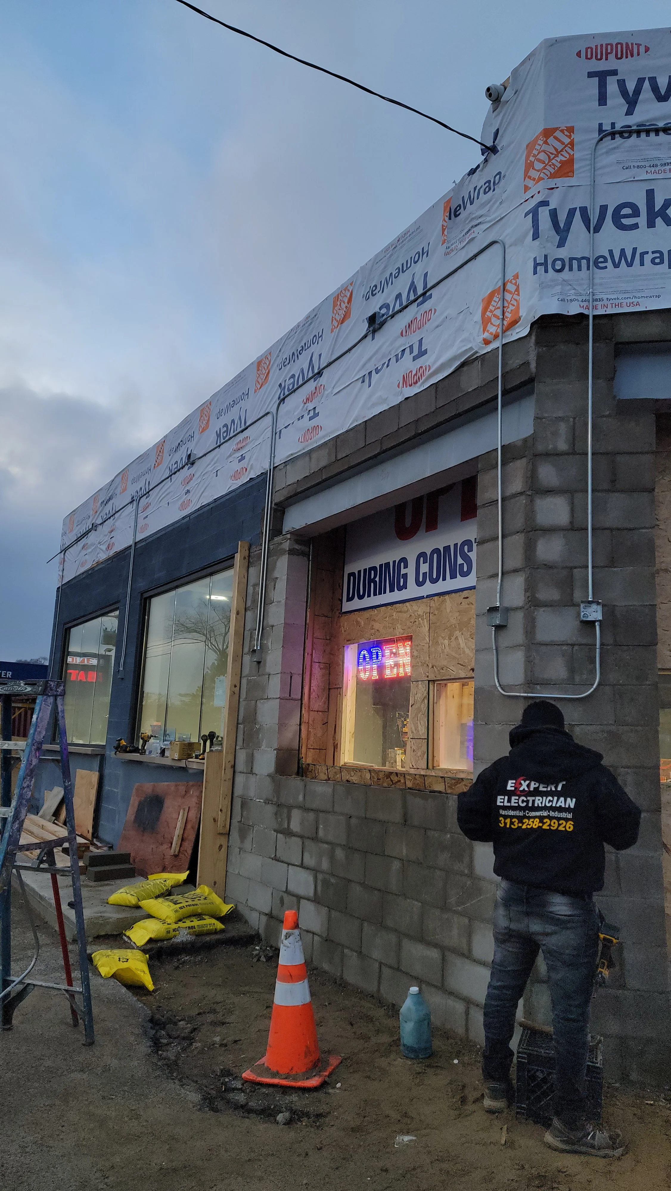 Construction worker standing outside a building under renovation, with bricks, tools, and construction materials visible. The building is partially covered with Tyvek HomeWrap and has an illuminated 'Open' sign in the window.