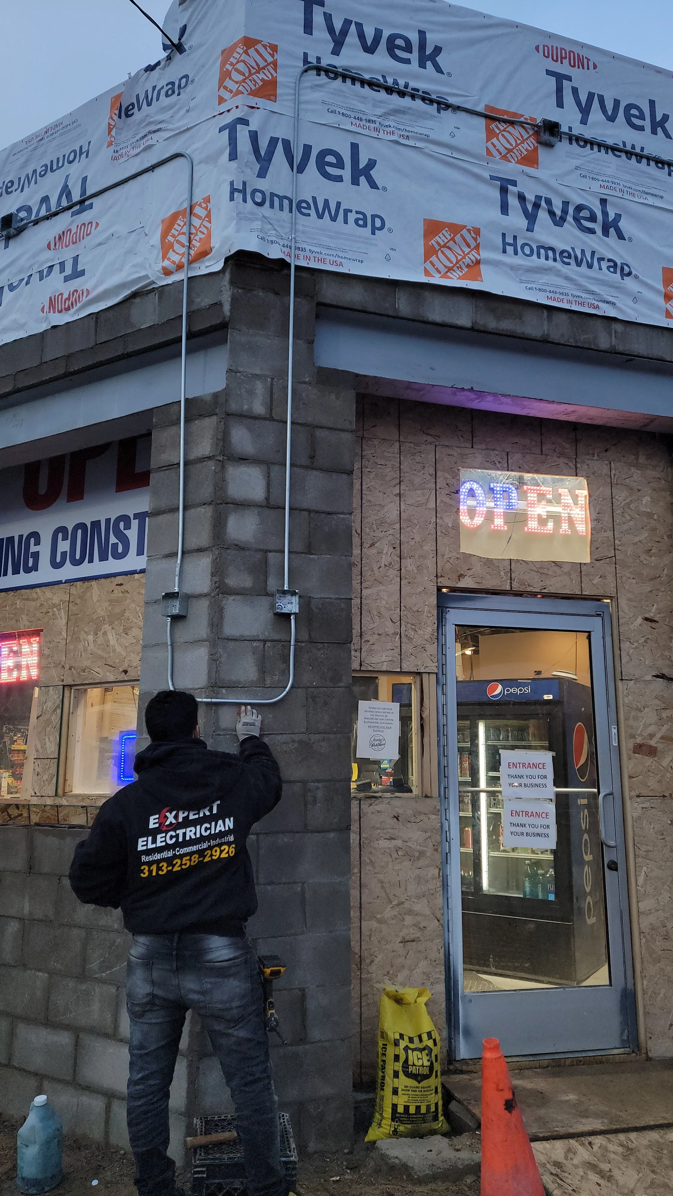 Worker installing electrical wiring on the exterior wall of a building under construction, with a neon 'OPEN' sign and a beverage store entrance in the background.