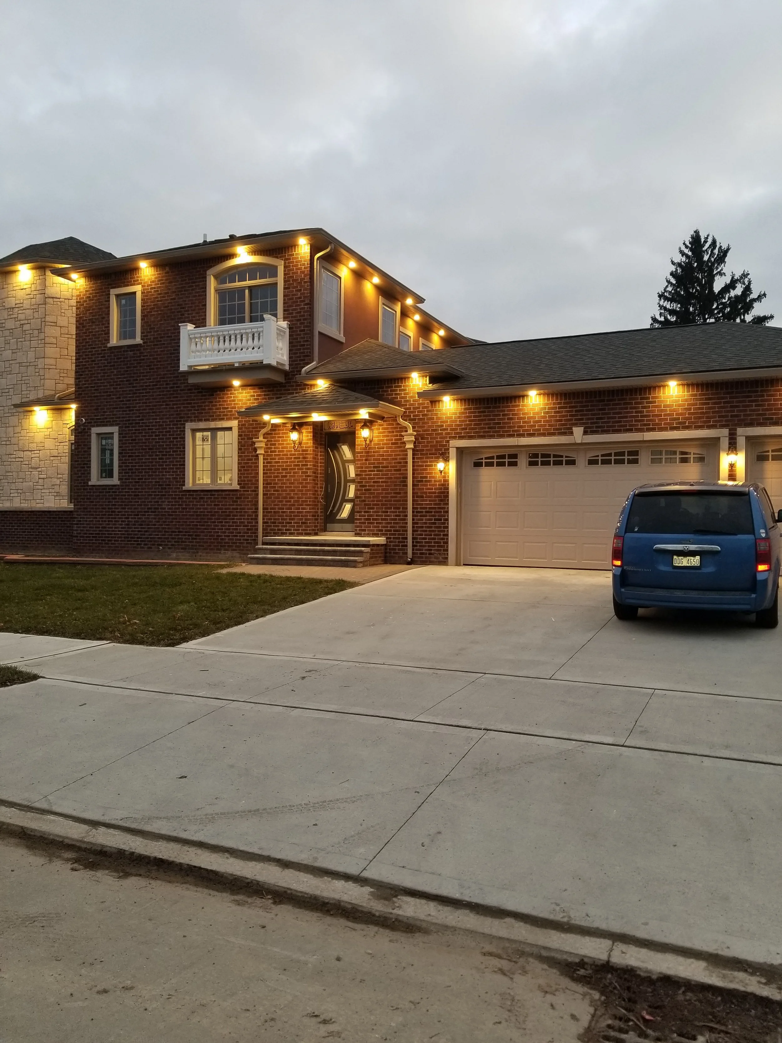 A two-story brick house with a stone and brick exterior, illuminated by exterior lights at dusk. The house features multiple windows, a small balcony, and attached garages. A blue vehicle is parked in the driveway.