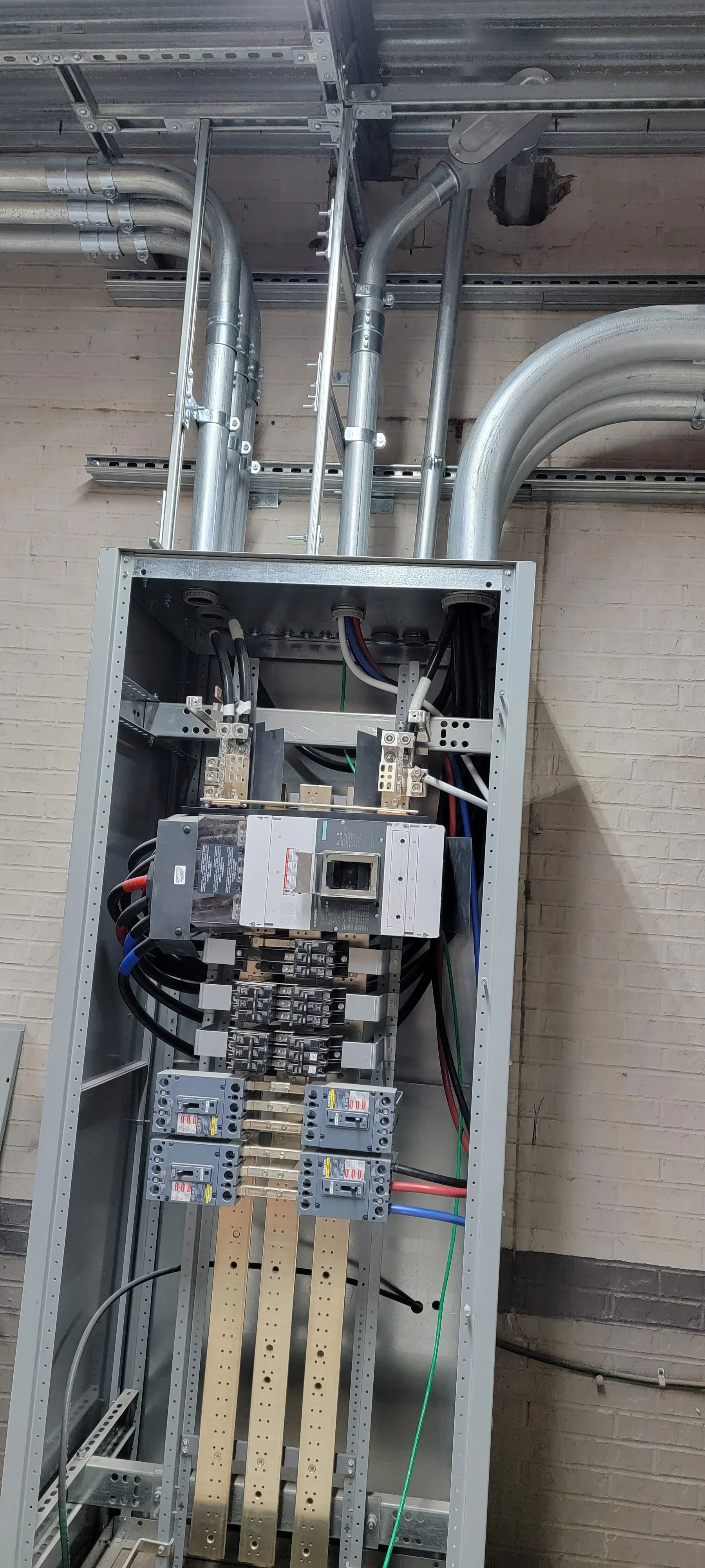 Electrical control panel with wiring, circuit breakers, and conduit pipes against brick wall.