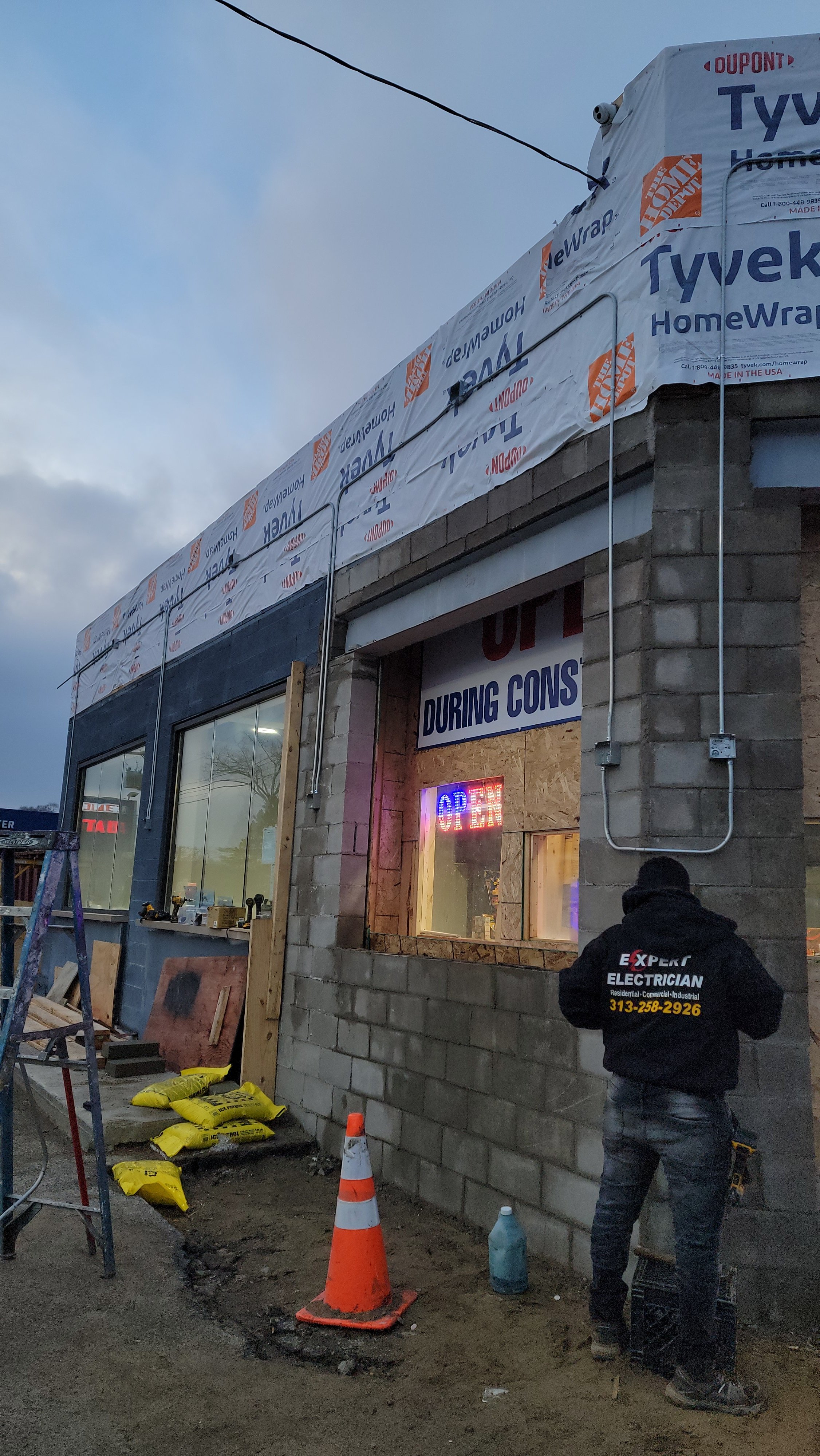 Construction site showing a building under renovation, with a worker in a black hoodie labeled 'Expert Electrician' on the back, construction materials, orange traffic cone, and a partially completed brick wall. The sky is cloudy, and there is construction wrap on the building's roof.