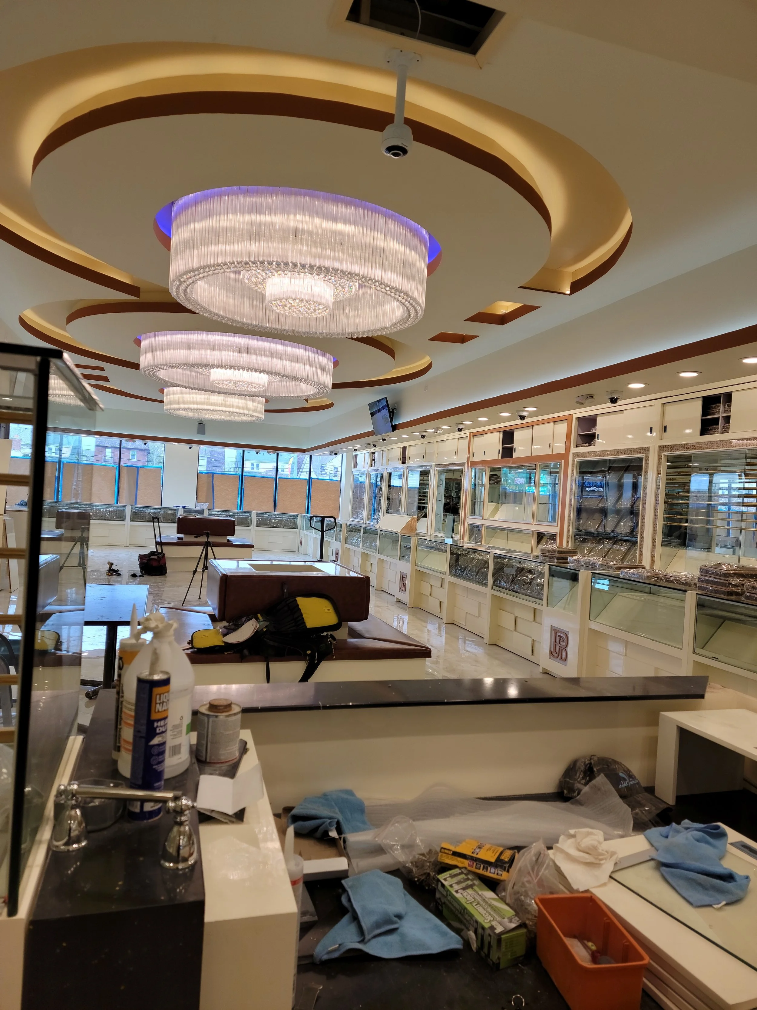 A restaurant or cafe interior with modern chandeliers, empty counters, equipment, and cleaning supplies scattered around. The room is well-lit with large windows at the back.