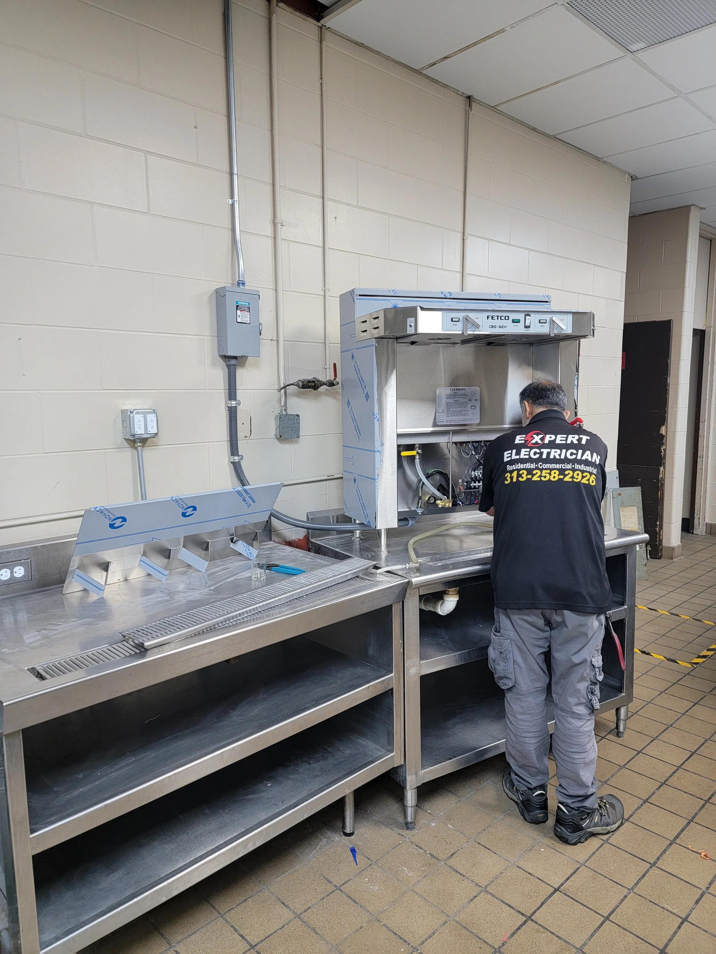 A worker in a black jacket with yellow phone number and the words 'Expert Electrician' is working on an electrical panel at a stainless steel work table in a commercial kitchen or industrial setting.
