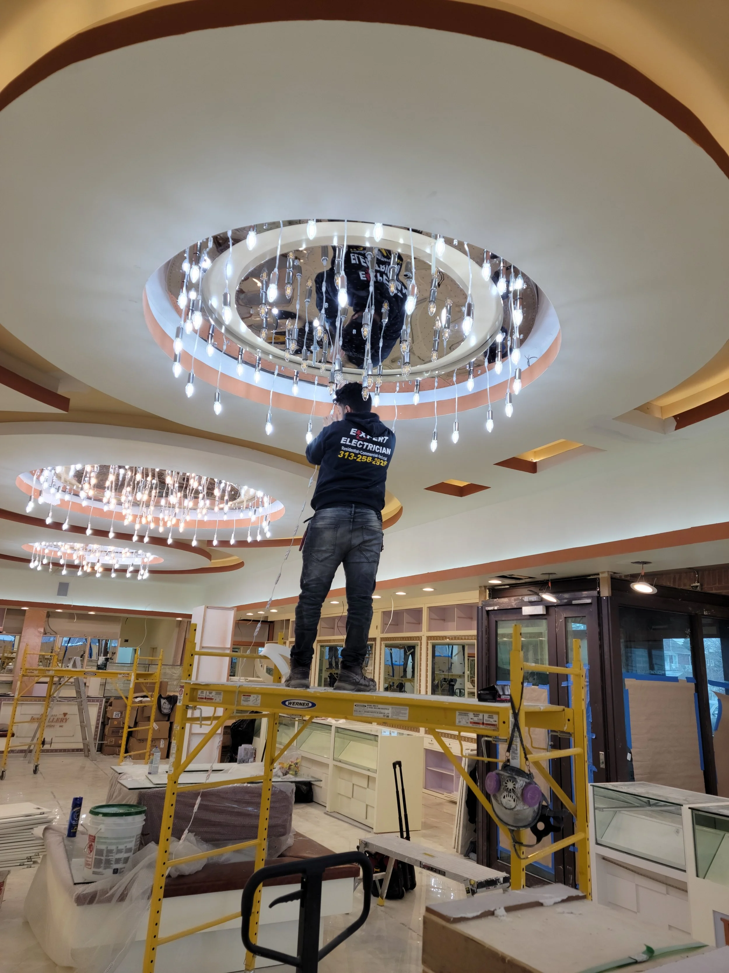 Two workers installing ceiling lights in a spacious indoor mall or store. One person stands on a yellow platform while the other is on a ladder, both working on the circular chandelier lighting fixture.