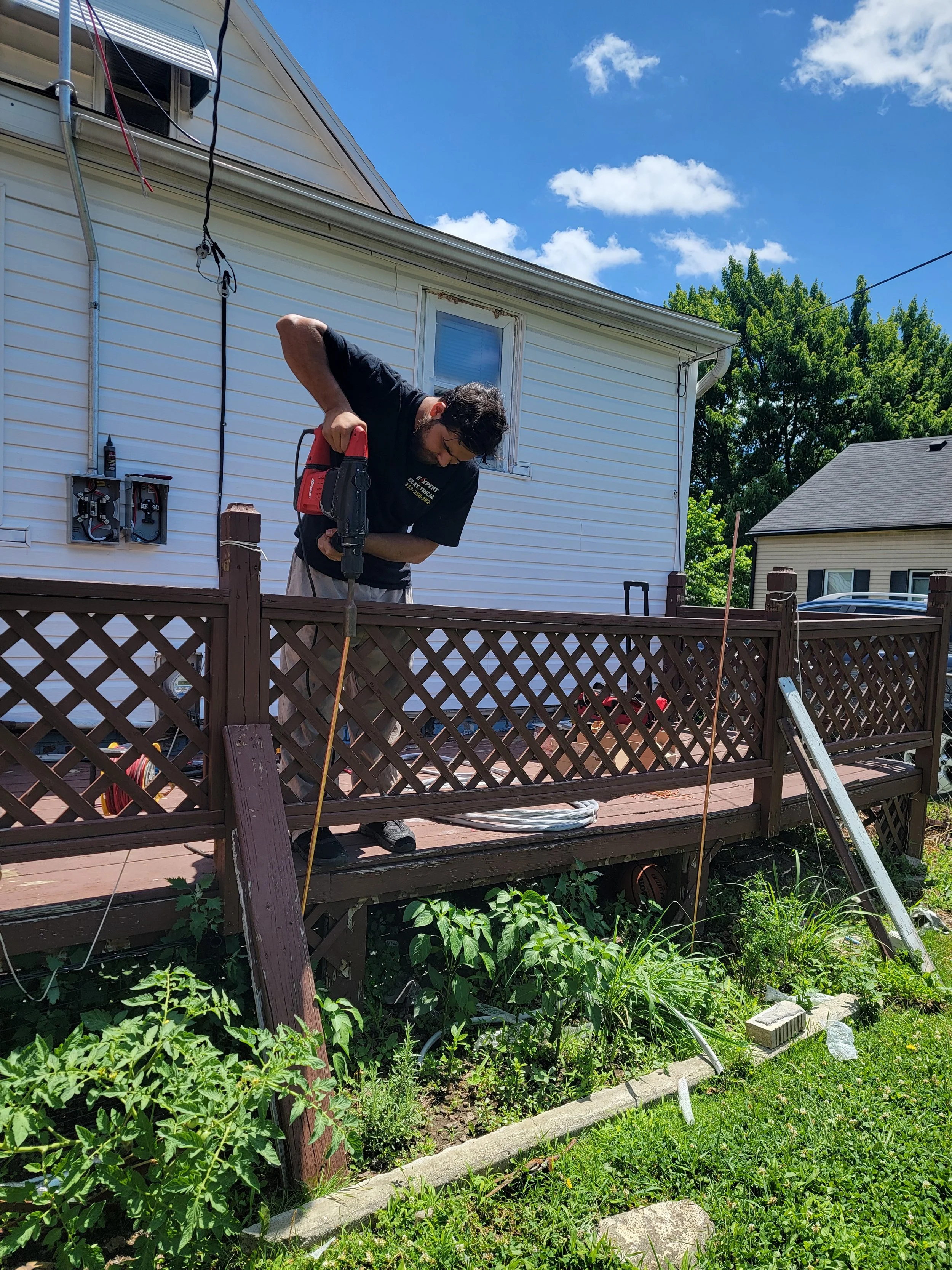 Man working outdoors in a backyard, using a power tool to build or repair a wooden deck, with greenery and a house in the background.