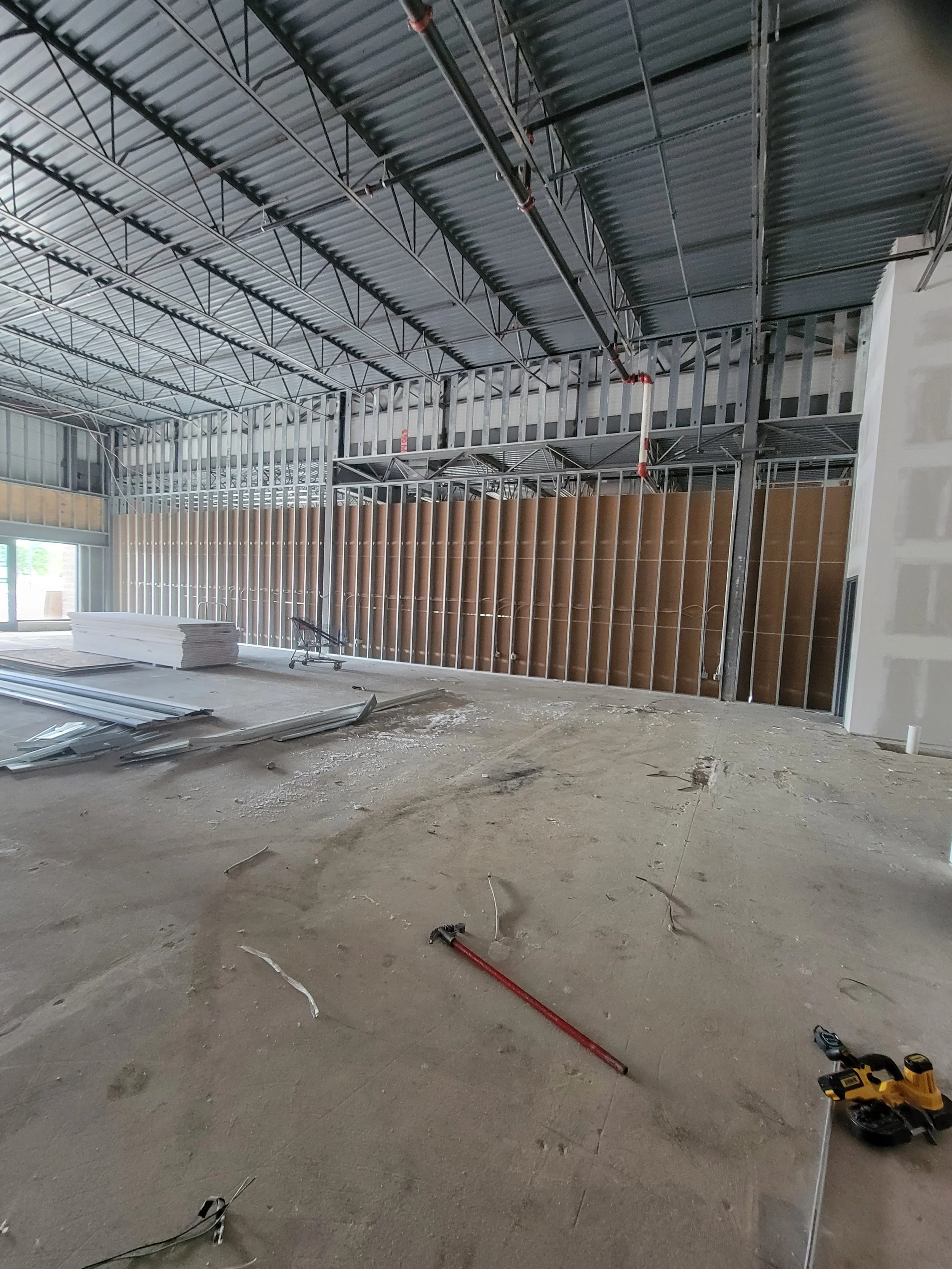 Interior view of a building under construction with exposed metal framing, electrical wiring, construction tools, and materials scattered on the unfinished floor.