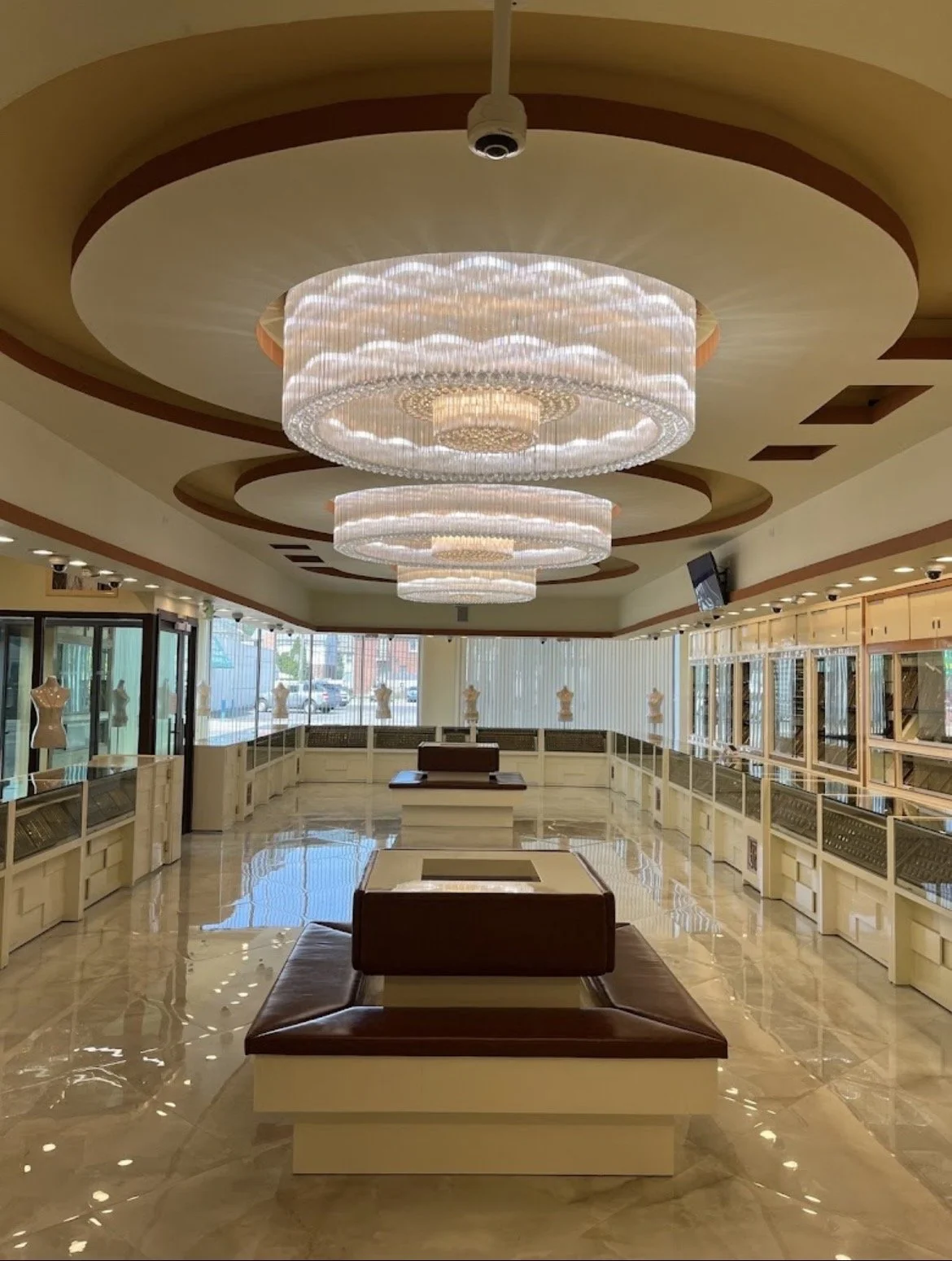 A spacious, elegant lobby with two large, circular chandeliers hanging from the ceiling. The room has polished marble floors, display mannequins on mannequins stands near windows, and a wooden seating area in the center. Large windows let in natural 