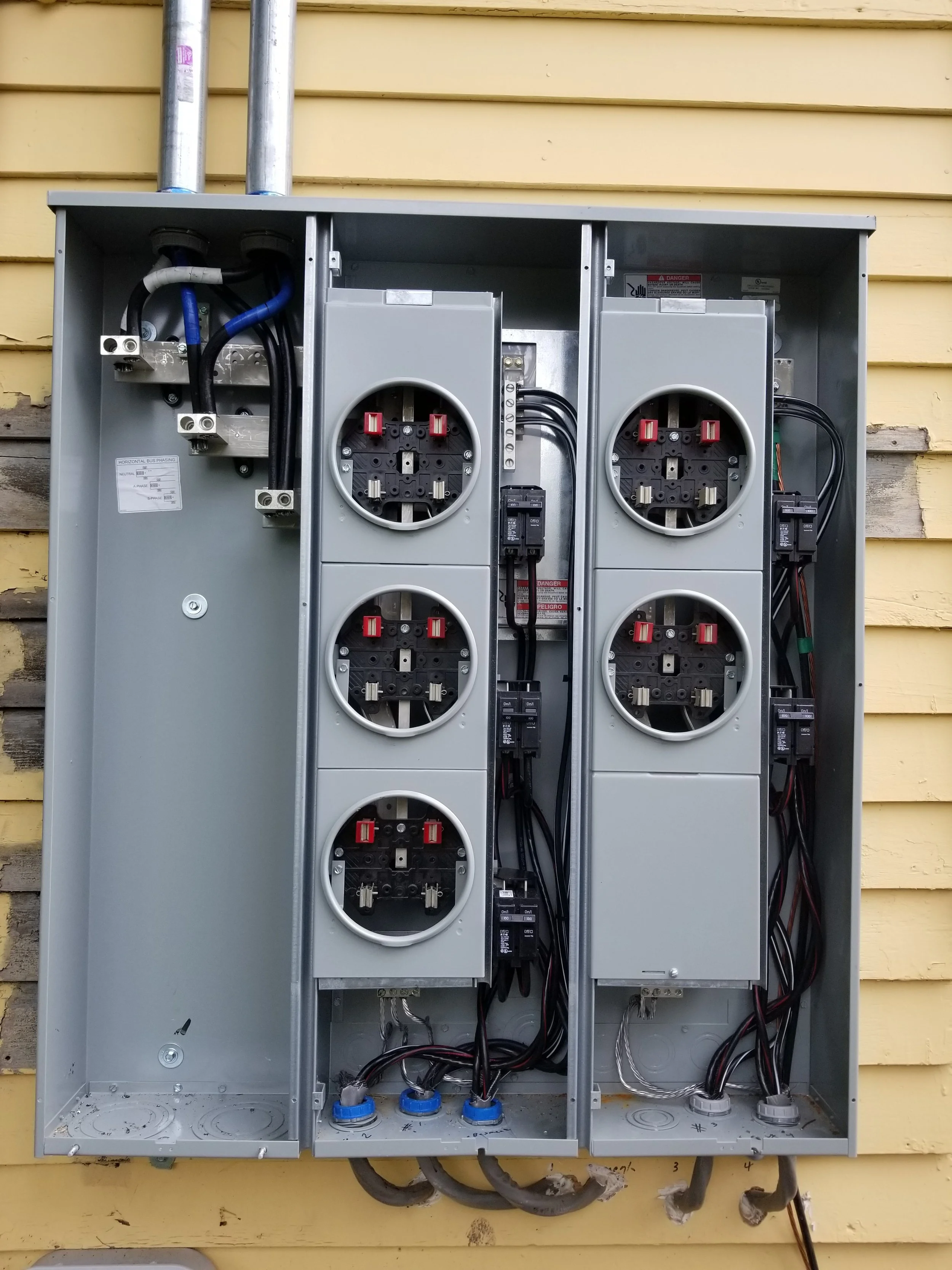 An open electrical meter box with wiring inside and no meters attached, mounted on a yellow wooden wall.