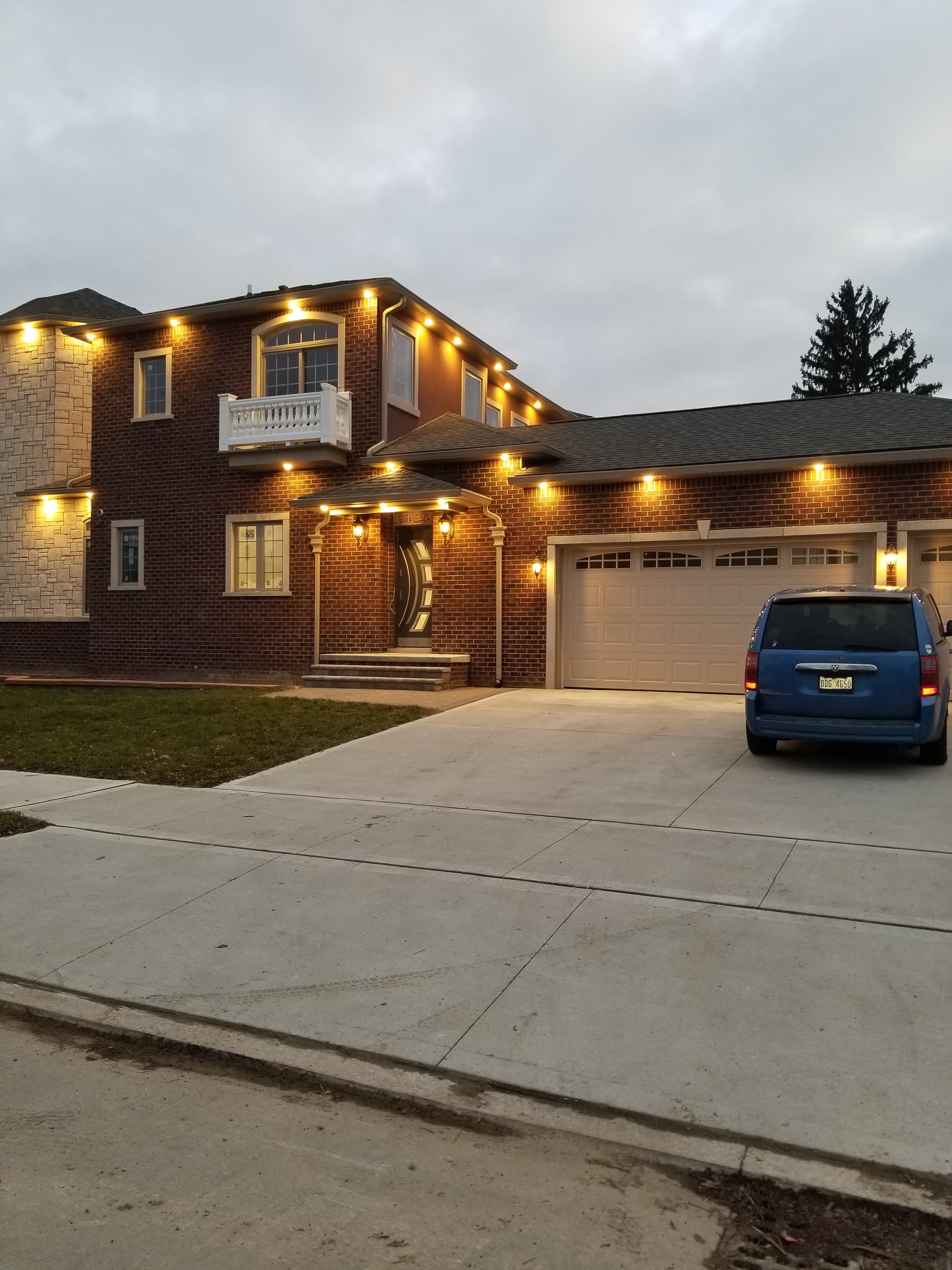 A brick house with outdoor lighting at dusk, with a small balcony on the second floor, a garage on the right, a blue minivan parked in the driveway, and a tree behind the house under a cloudy sky.
