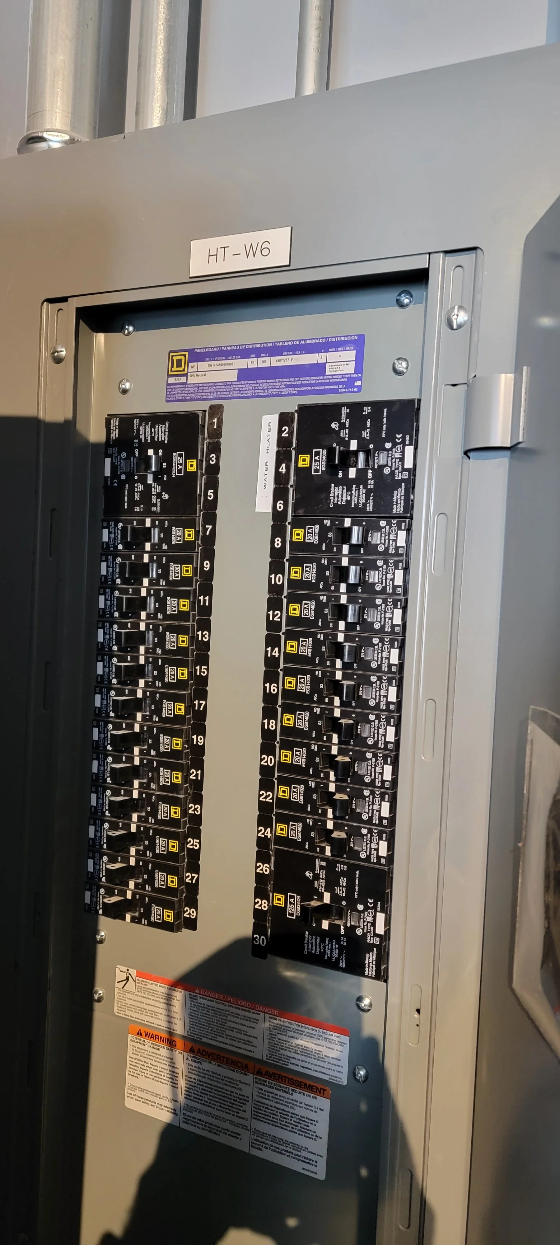 An electrical breaker panel with multiple black switches, labeled 1 through 30, organized in two columns, with a sticker indicating "Water Heater" and a small purple label at the top. The panel has a metal door with a label "HT-W6" and warning labels
