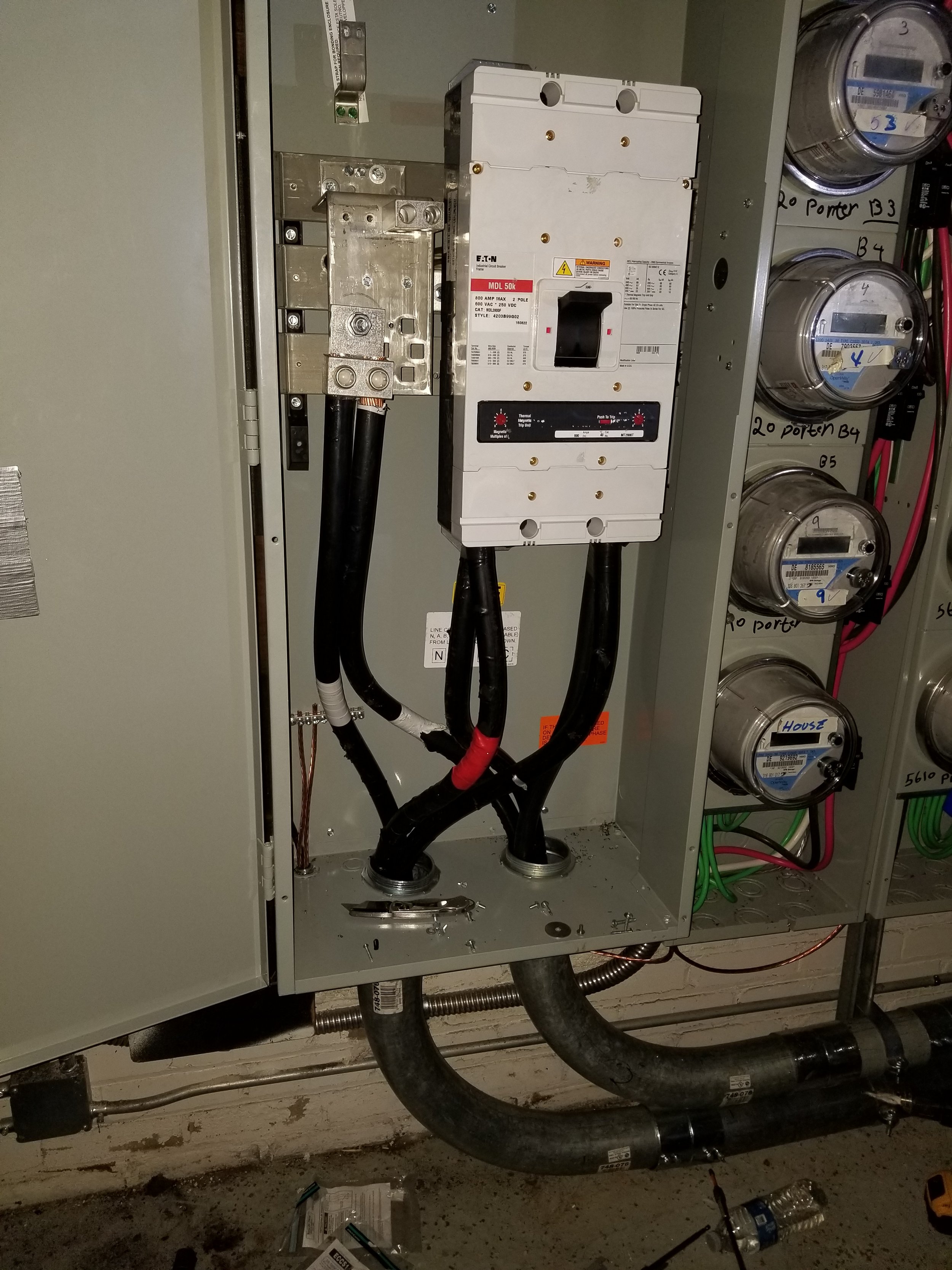 Close-up of an electrical panel with wires, circuit breaker, and dials, partly open and under maintenance, with tools and debris on the floor.