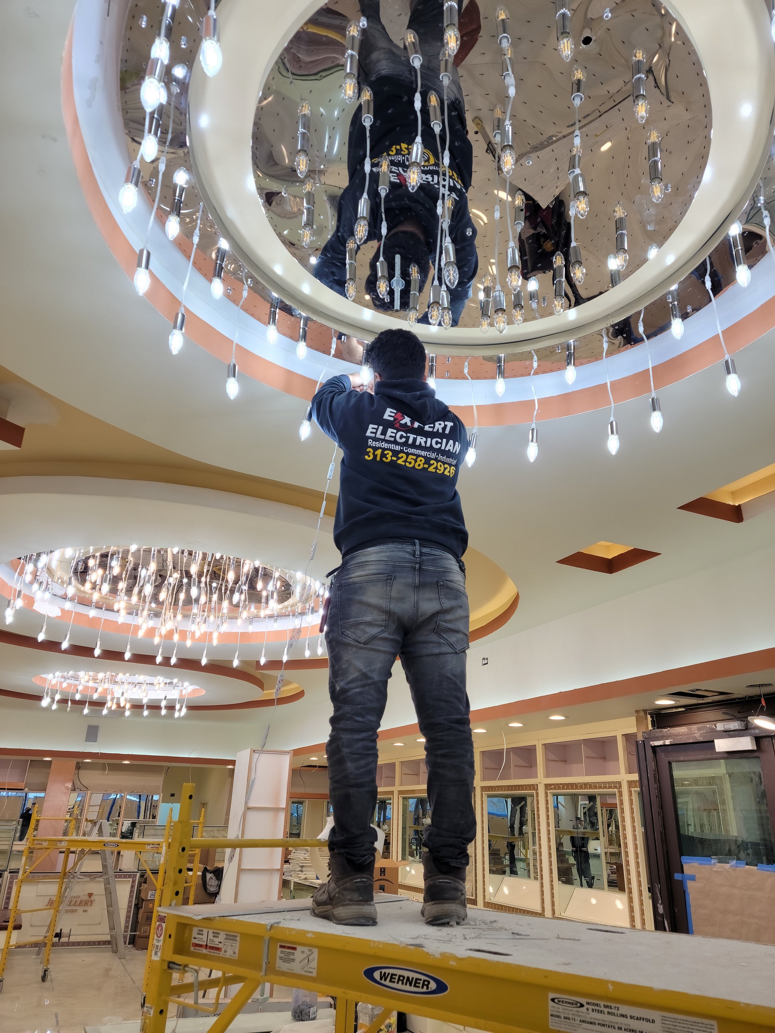 A worker installing or working on a ceiling fixture in a large, well-lit building, using a scaffold table with the brand 'WERNER'. The ceiling has circular, decorative lighting with hanging bulbs.