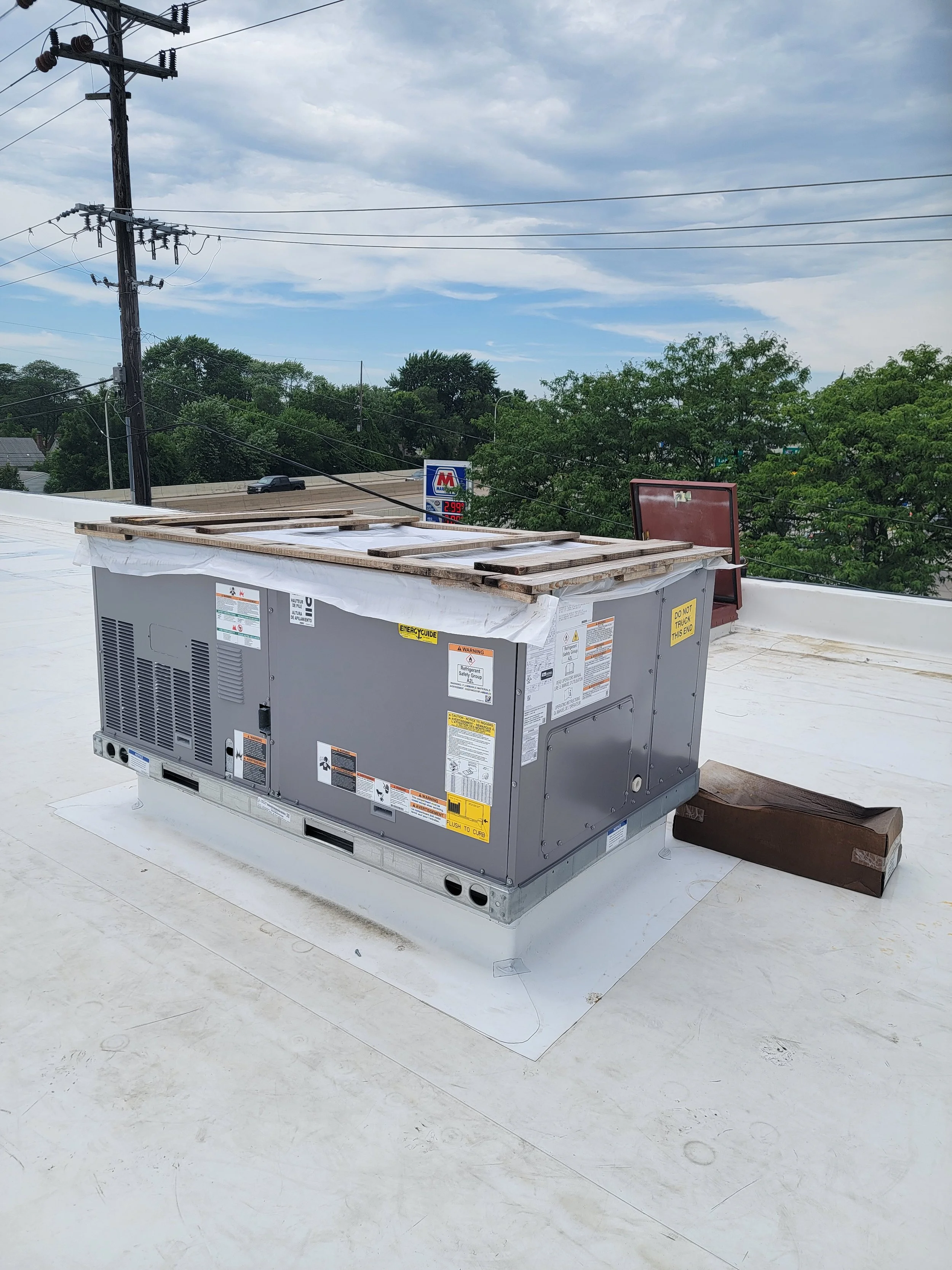 HVAC rooftop unit on a white roof with wooden pallets and utility poles with power lines, trees, and a sign for a convenience store in the background under cloudy sky.