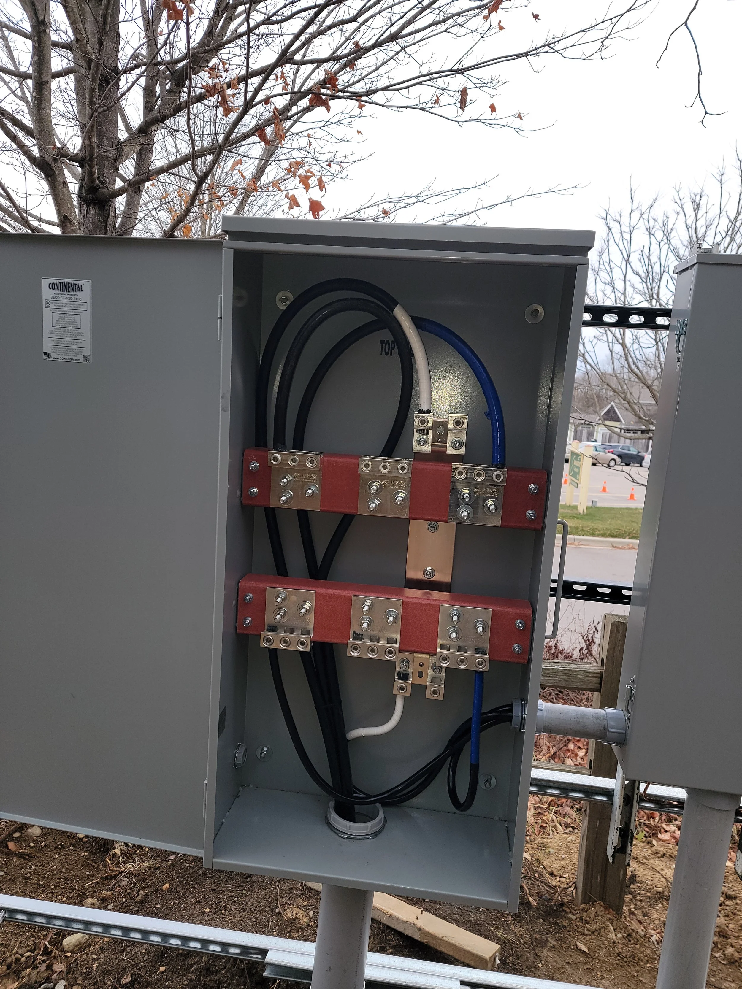 An opened gray metal electrical meter box with internal wiring and circuit breakers, with a background of leafless trees and a suburban neighborhood.