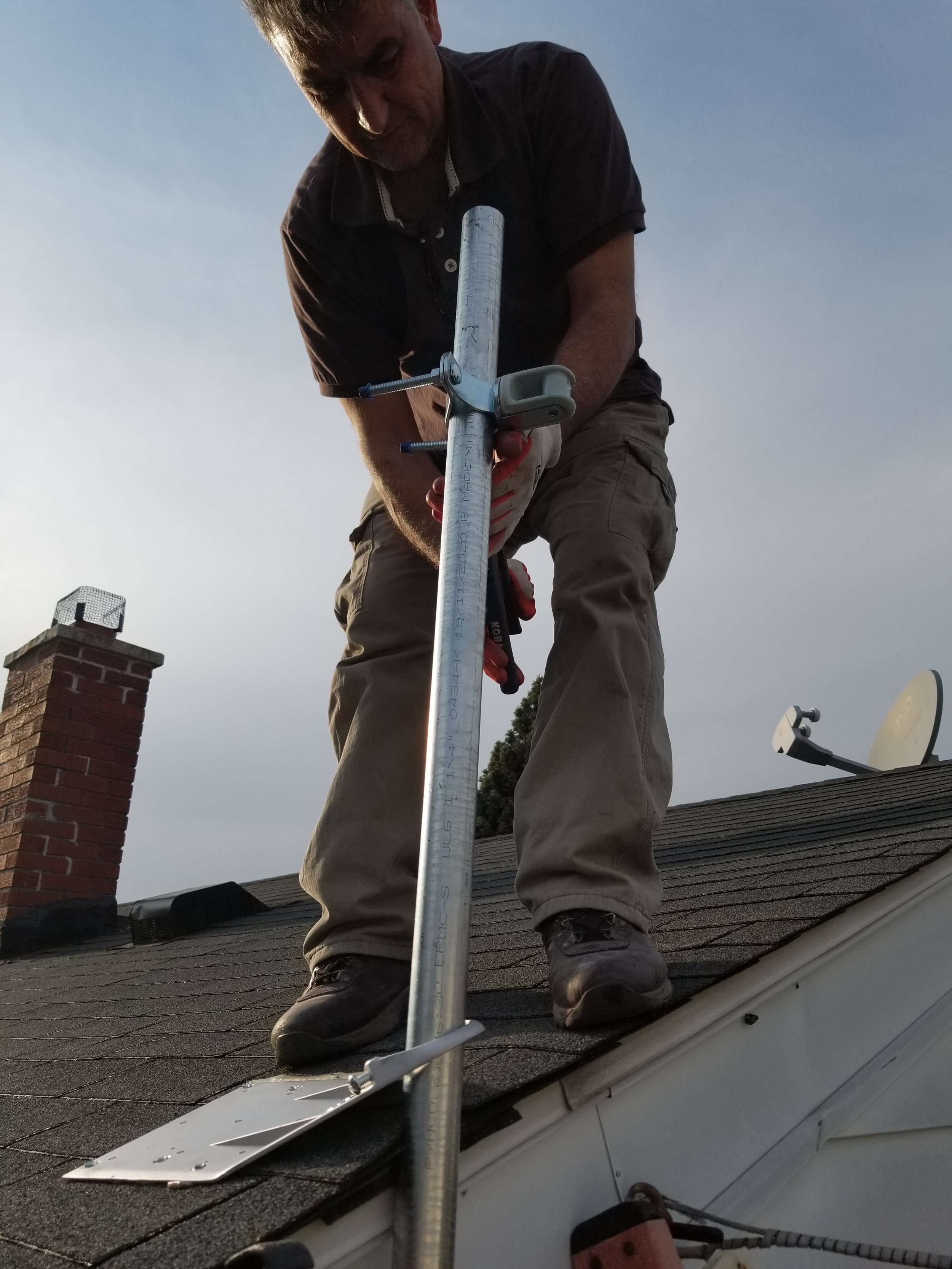 A man working on a roof, using a pole with a measurement tool attached, with a chimney and satellite dish visible in the background.