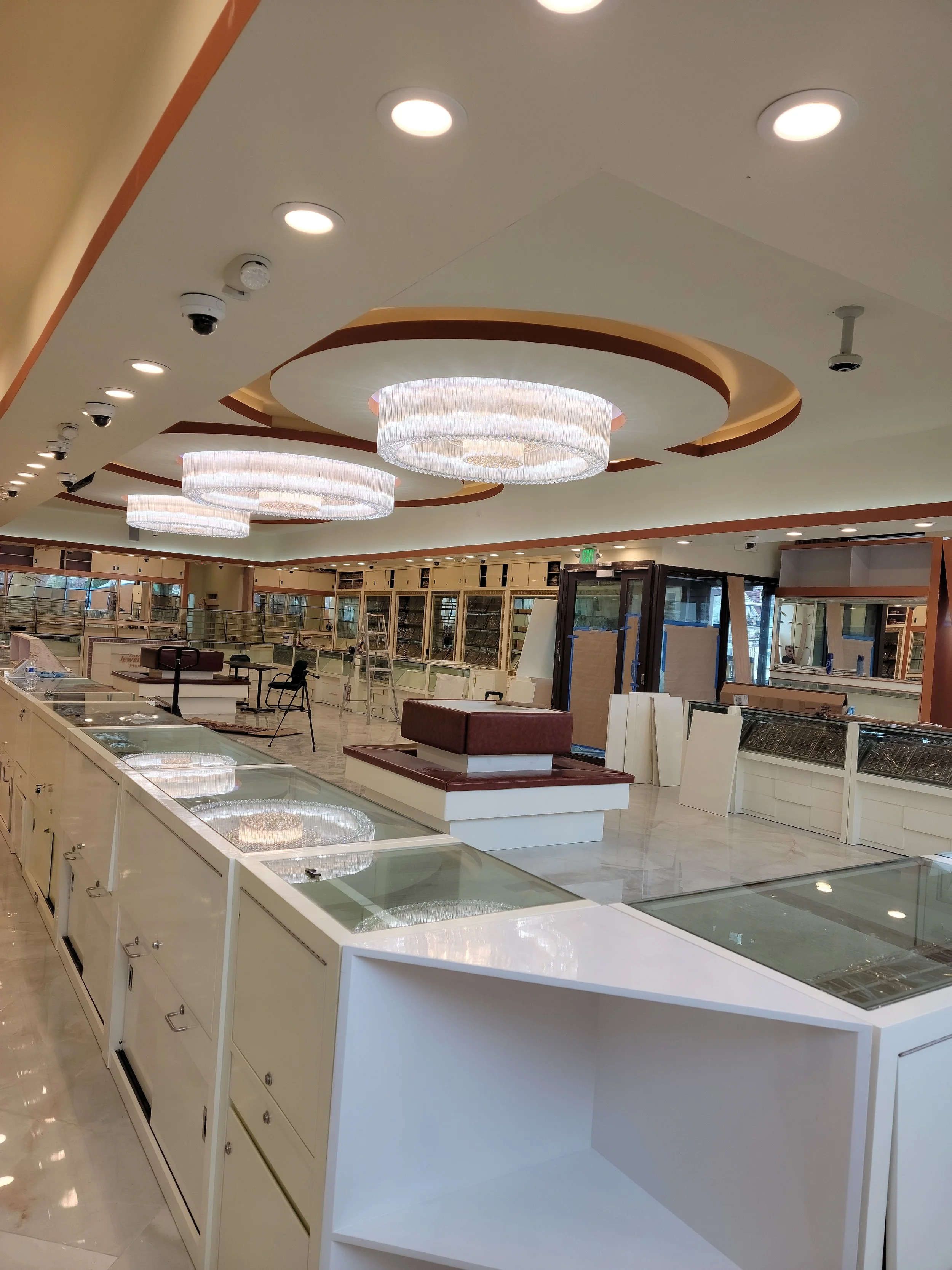Interior of a retail store or jewelry shop under renovation, with display counters, ceiling lights, chandeliers, and construction materials like ladders and chairs.