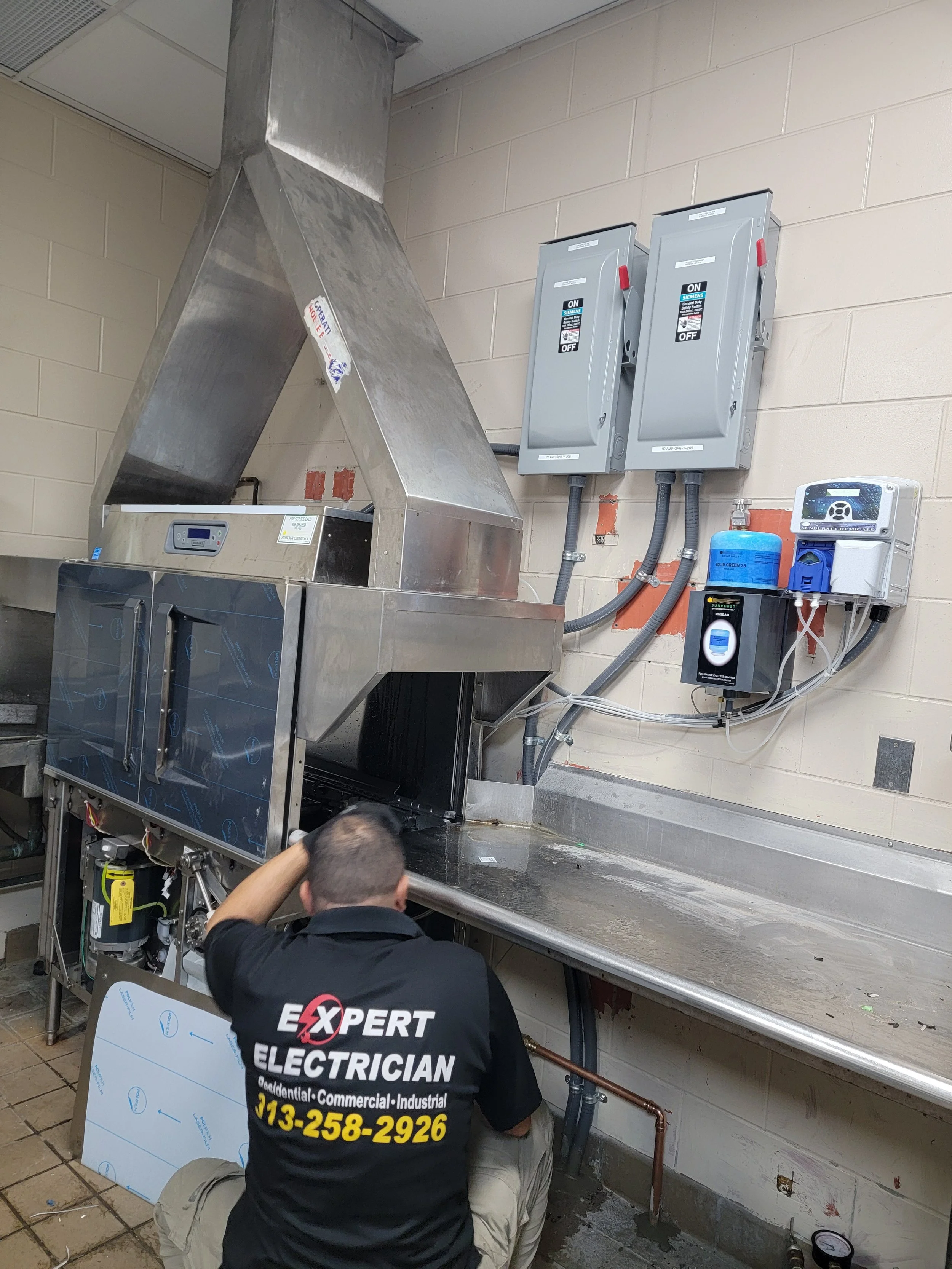 A technician working on electrical or industrial equipment in a commercial kitchen or industrial setting, with electrical panels and pipes on a beige brick wall in the background.