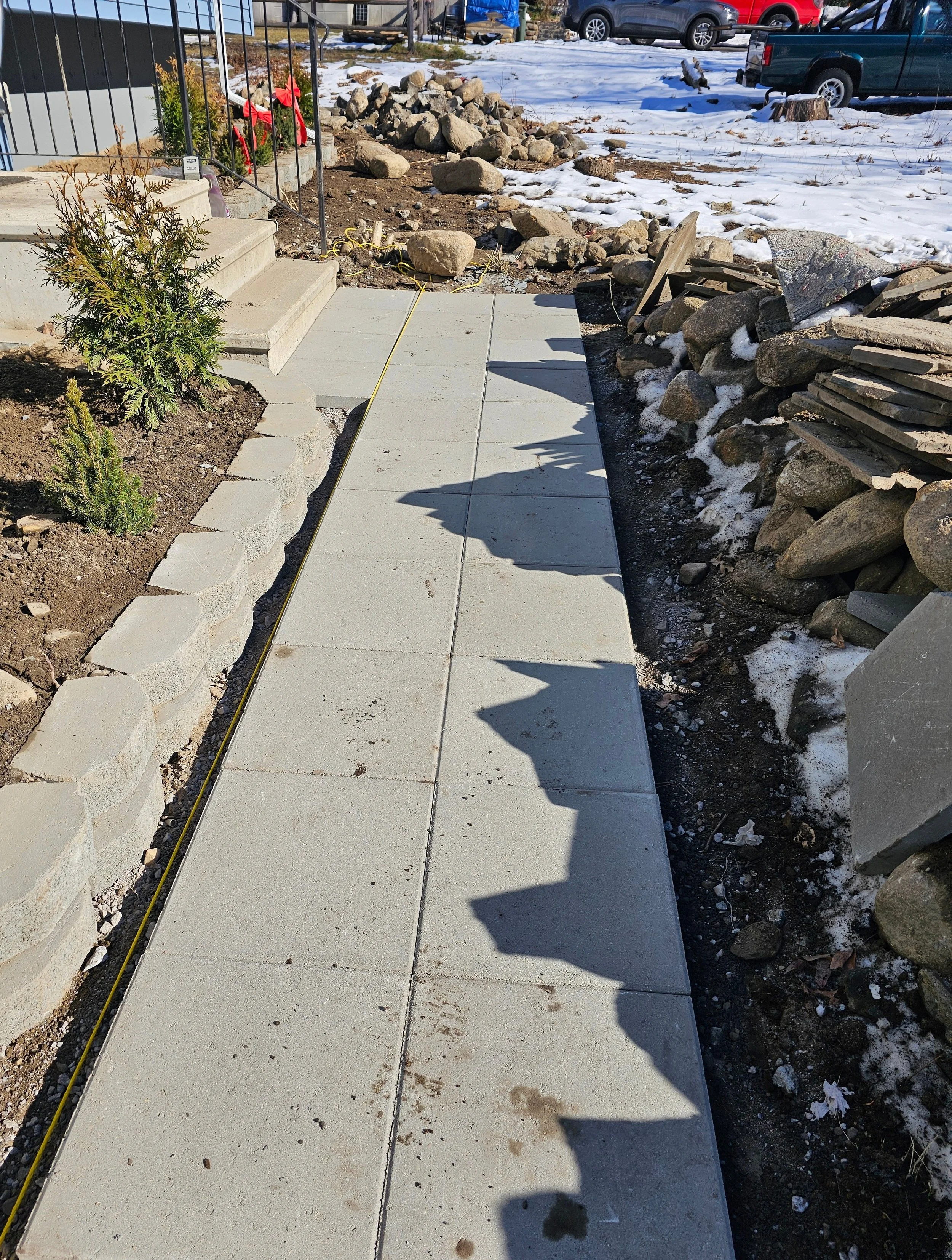 A sidewalk under construction or repair, with orange construction tape and yellow measuring tape, showing new concrete slabs, rocks, and a gravel area with some snow in the background.