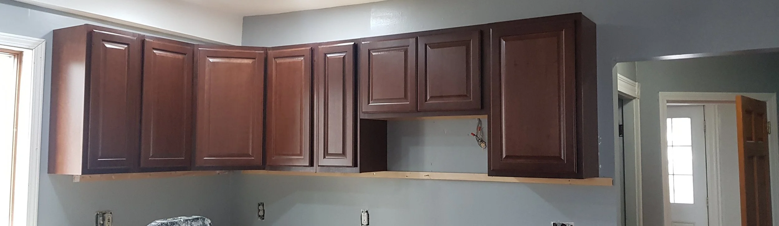 Kitchen cabinets being installed, with some electrical wiring visible and an open space for a microwave or oven, in a room with gray walls.