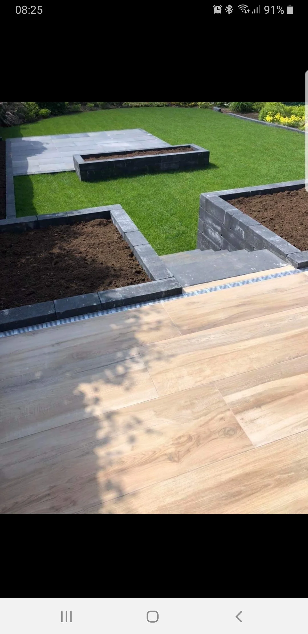 A backyard with a newly installed wooden deck, a brick garden bed, and a well-maintained lawn with some paving stones in the distance.