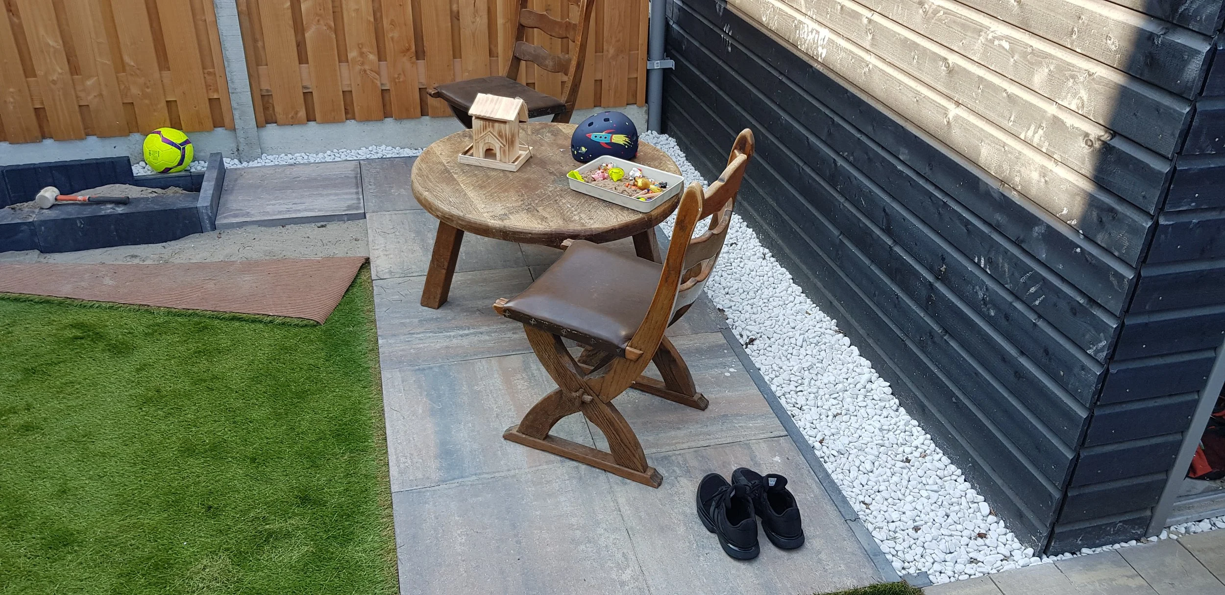 Backyard patio with a small round wooden table and two chairs, one with a pair of black shoes underneath. Items on the table include a wooden birdhouse, a black helmet with a colorful rocket design, and a white tray with small toys. To the left, a partially filled sandbox with a tool inside, a bright green soccer ball, and a mat on artificial green grass. The backyard is enclosed with a wooden and black slatted fence, with gravel along the edges.