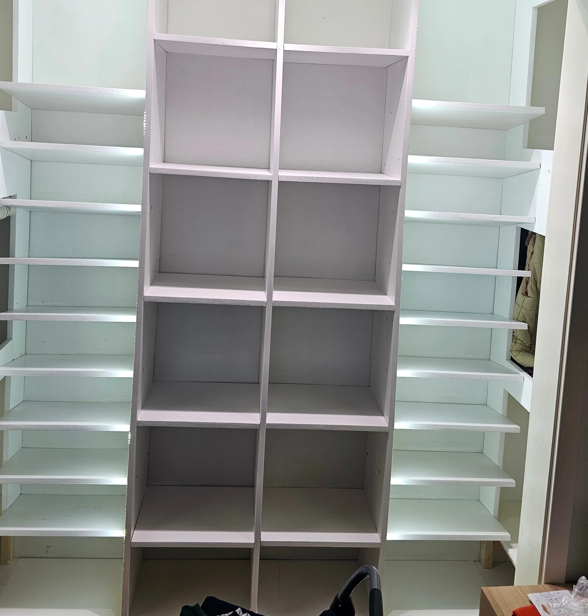 Empty white wooden cubby shelves in a room, with additional shelves in the background and various items in the lower part of the image.