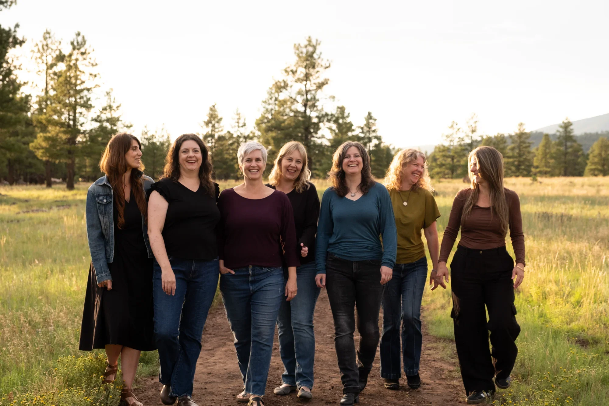 Six women walking together outdoors on a trail in a forested area, smiling and talking to each other during sunset.