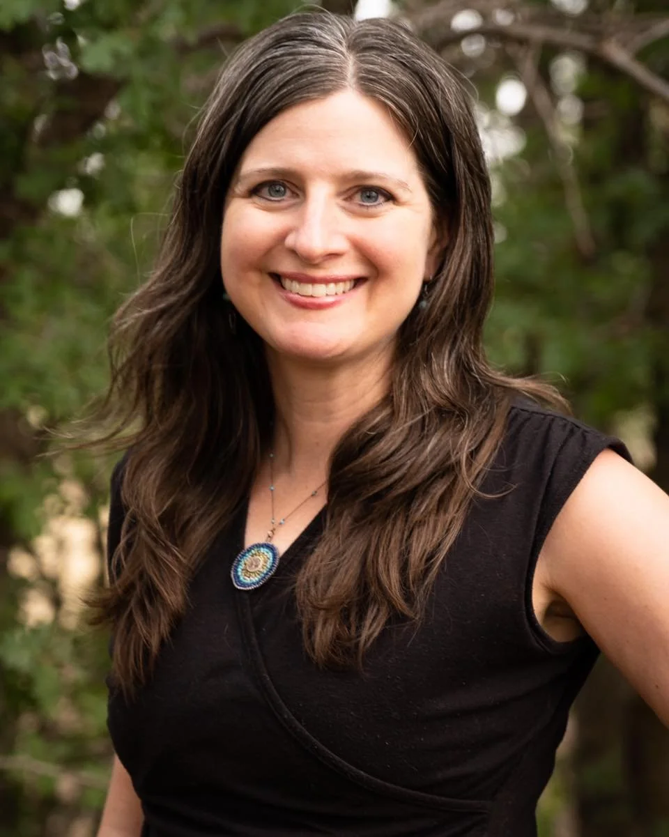 A woman with long brown hair and blue eyes smiling outdoors, wearing a black sleeveless top and a colorful necklace, with green foliage in the background.