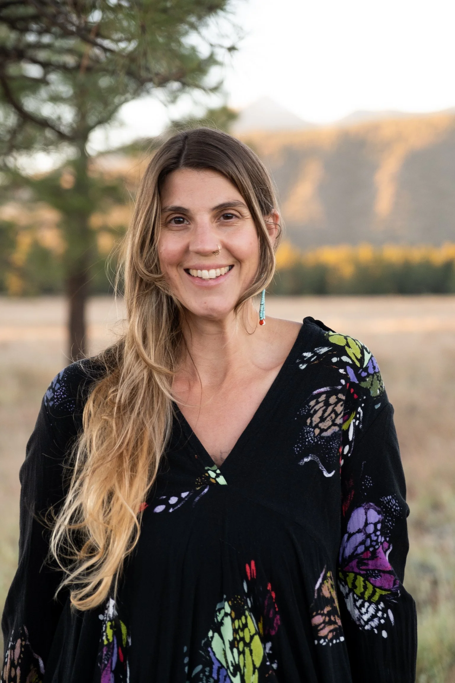 A woman with long wavy hair smiling outdoors, wearing a black top with colorful butterfly and floral patterns, with trees and mountains in the background.