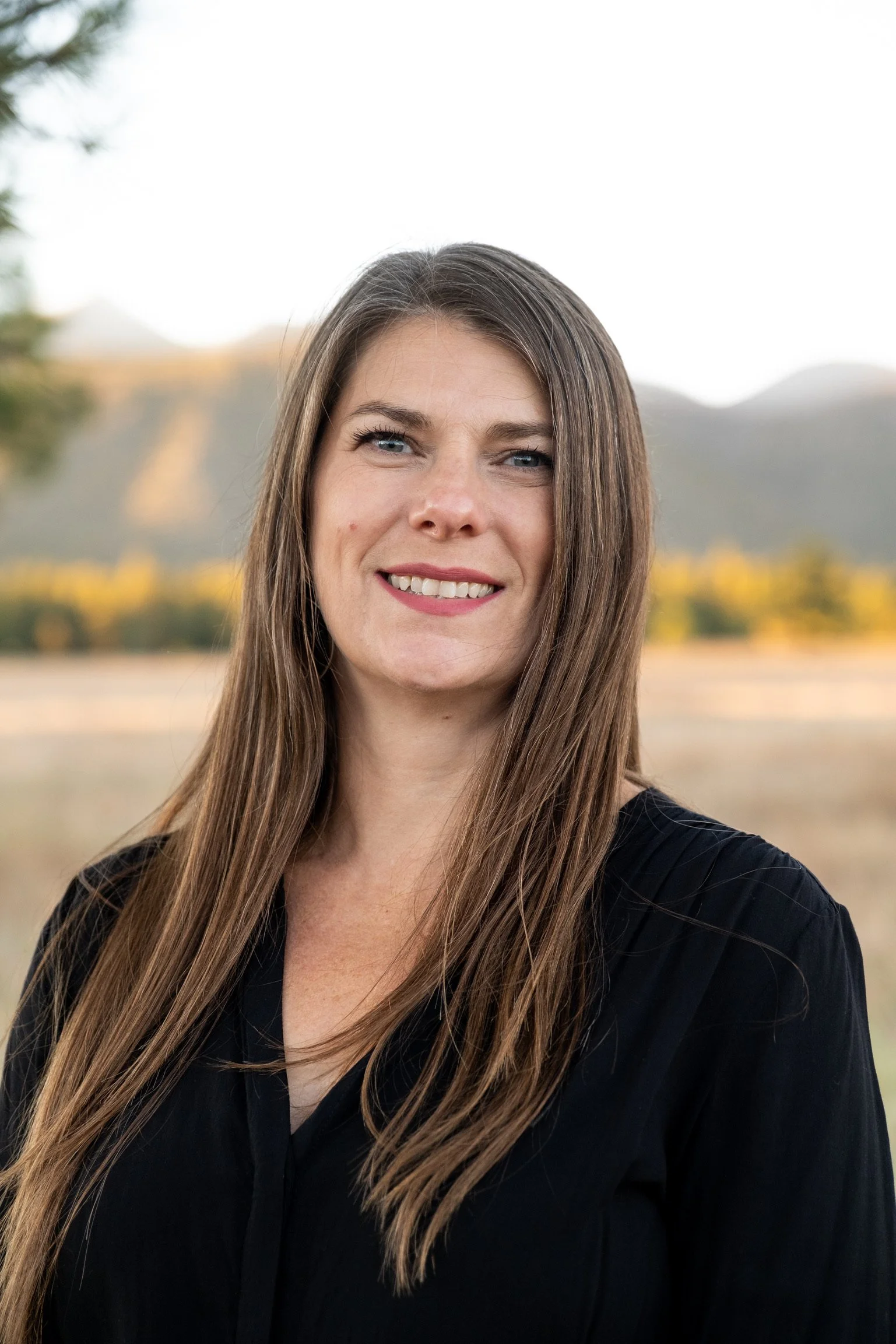 A smiling woman with long brown hair wearing a black top outdoors with a blurred landscape of mountains and trees in the background.