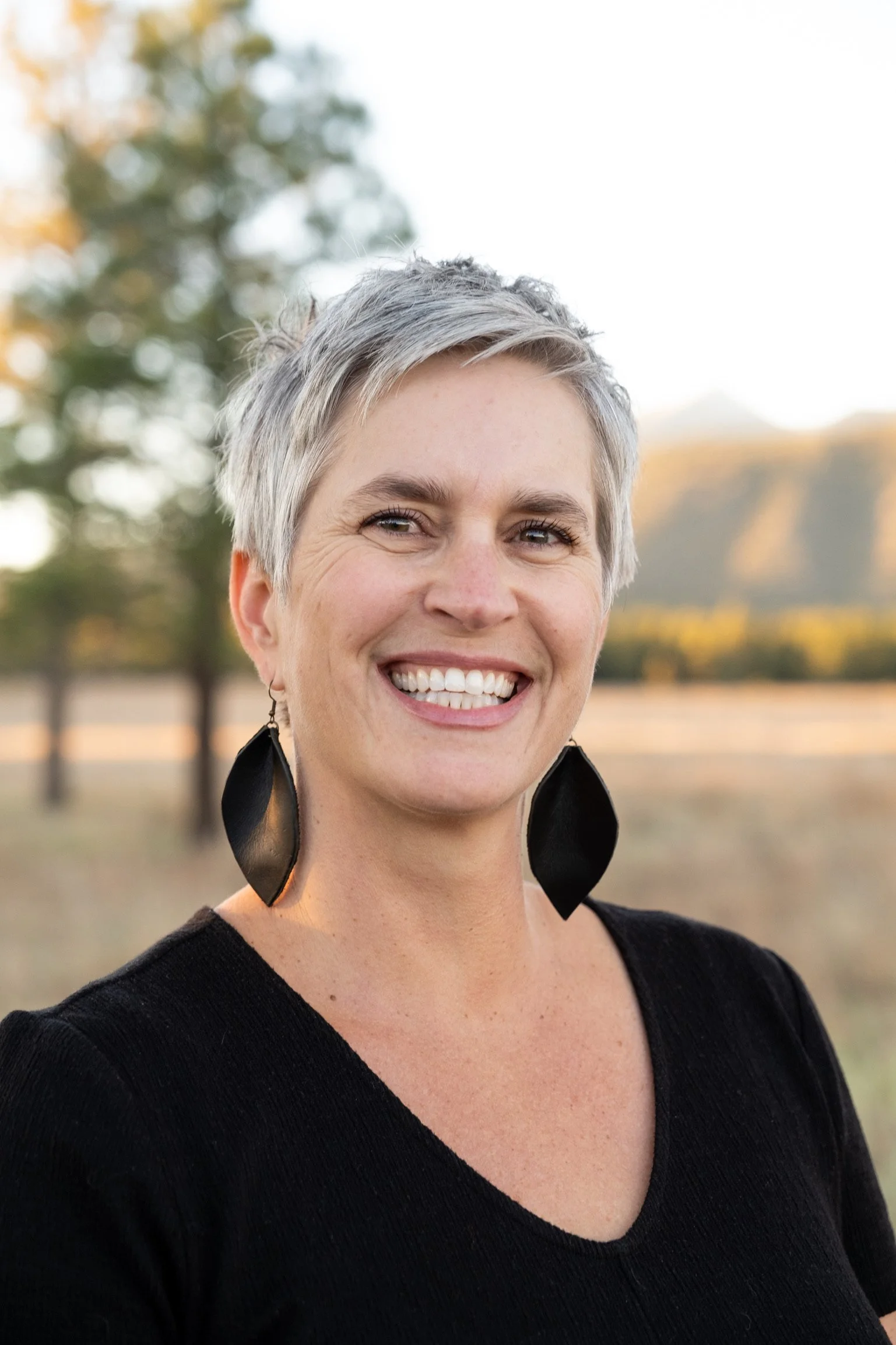 Close-up of smiling woman with short gray hair, wearing large black earrings and black top, outdoors with blurred trees and sky in the background.